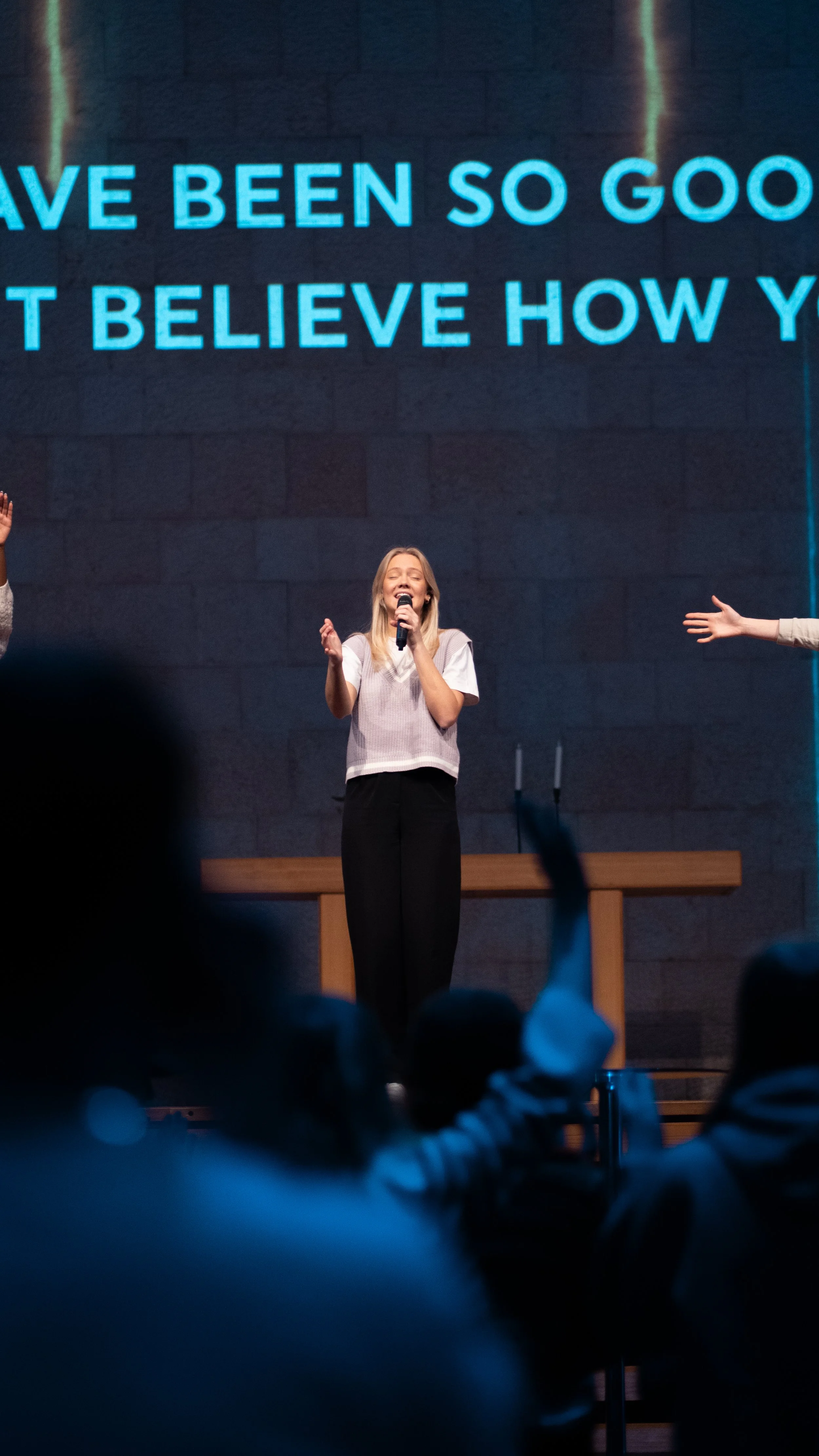 A woman singing on stage with a large screen behind her displaying partial song lyrics in blue text.
