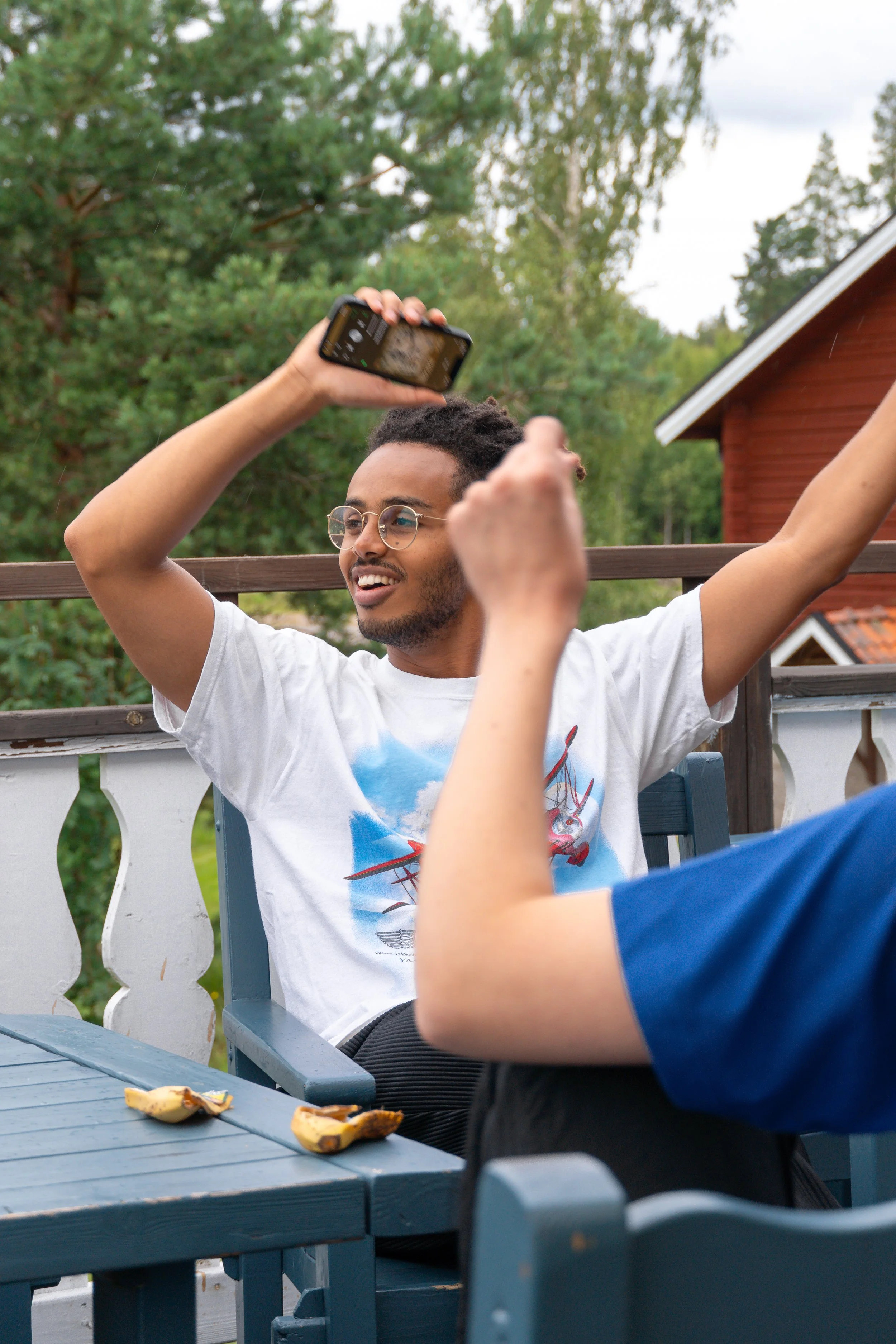 A young man wearing glasses and a white t-shirt with a graphic print is sitting outdoors on a patio, holding a smartphone above his head, taking a photo or video. Another person is partially visible in front of him, and there are banana peels on the blue table. In the background, there are trees and a red house.