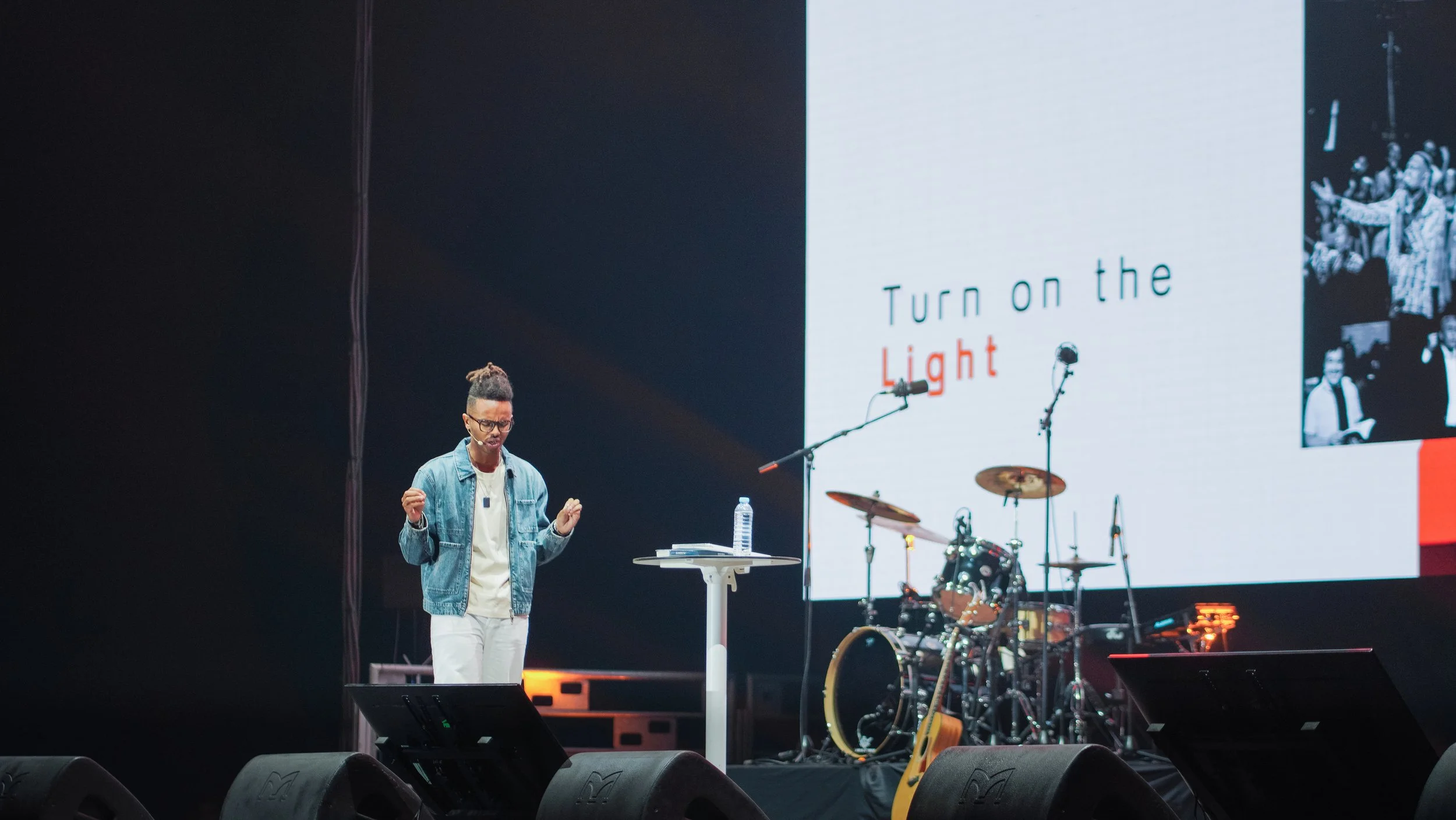 A person speaking on stage with a large screen behind them displaying the text "Turn on the Light." The stage has musical instruments including a drum set and an acoustic guitar.
