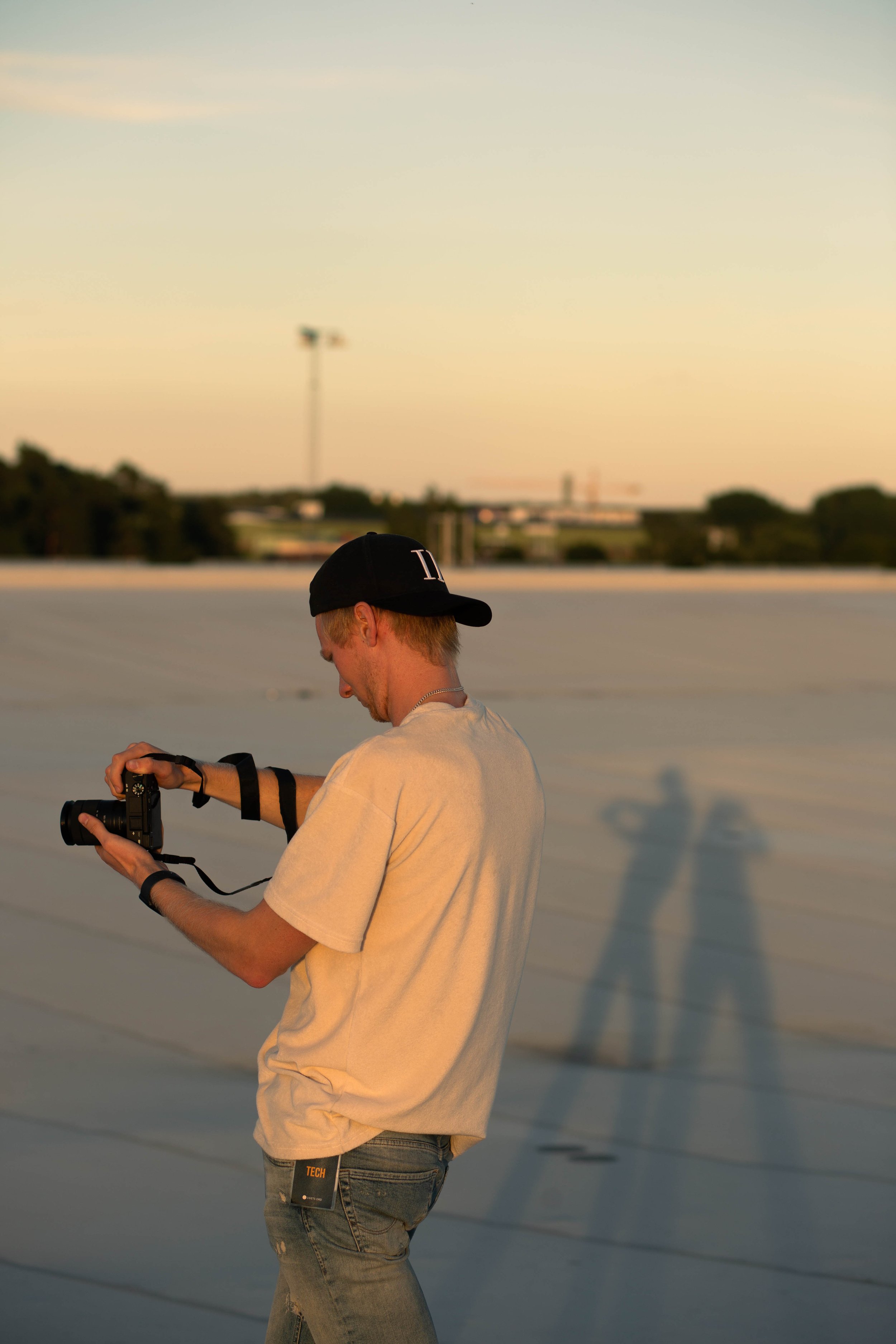 A young man standing on a rooftop during sunset, looking at his camera. His shadow is cast on the roof, with the sunset sky in the background.
