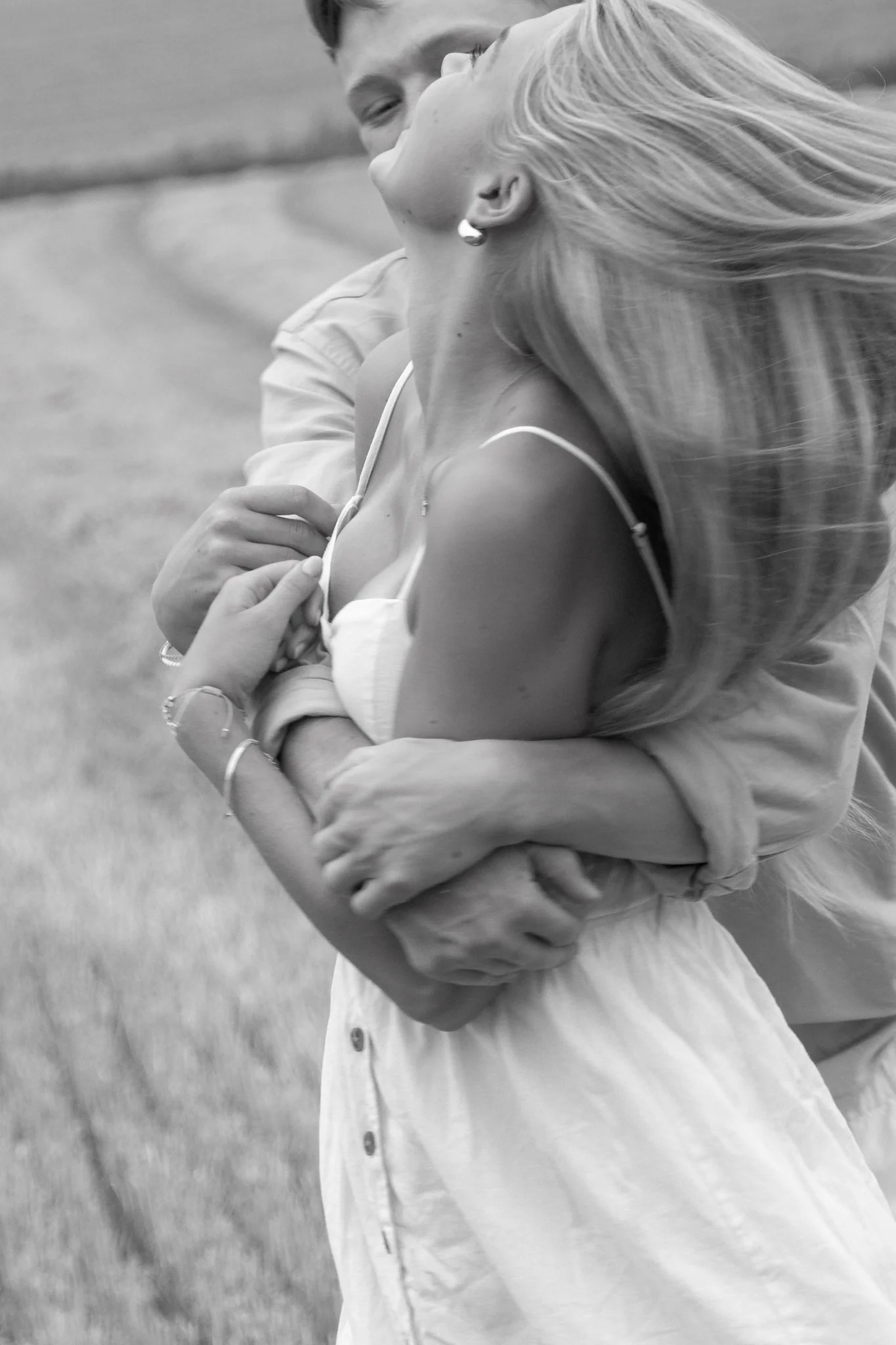 A black and white photo of a couple embracing, with the woman slightly leaning back and the man holding her from behind. The woman's long hair is flowing, and she is wearing a sleeveless top and jewelry.