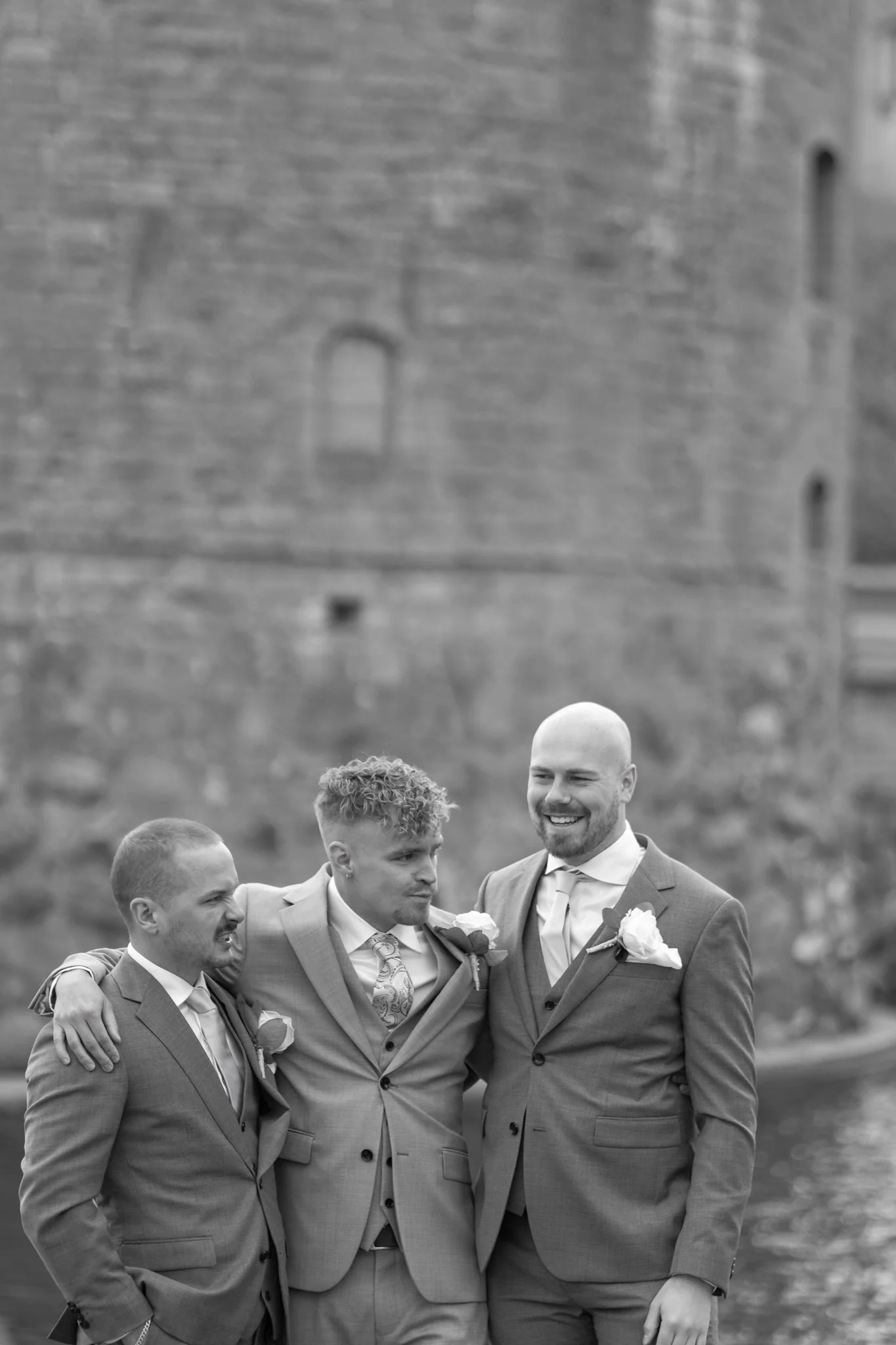 Three men in suits at a wedding, embracing and smiling outdoors with a blurred stone building in the background.