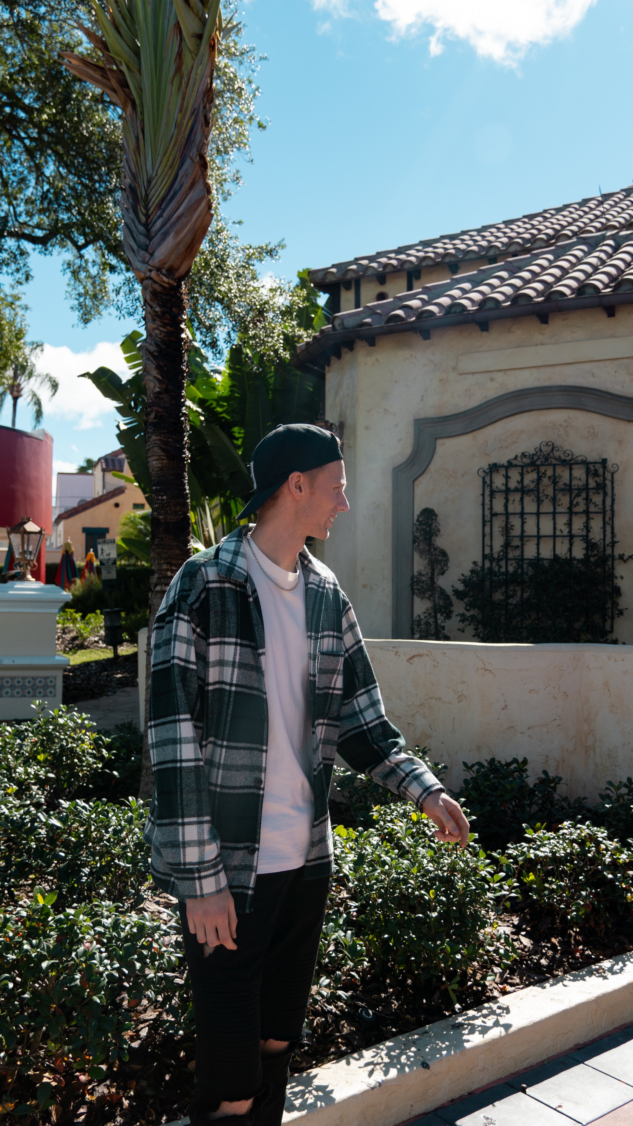 A young man wearing a backwards baseball cap, white t-shirt, plaid jacket, and black ripped jeans, standing outside in a sunny area with greenery and Mediterranean-style buildings in the background within Universal Studios in Florida, USA.