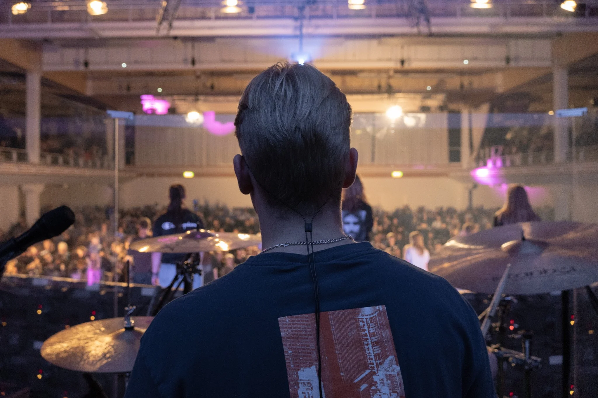 A person with blonde hair viewed from behind, standing on stage at a concert or event with a large crowd in front of him. The stage features drums and microphone equipment, with colorful lighting and a two-story venue.
