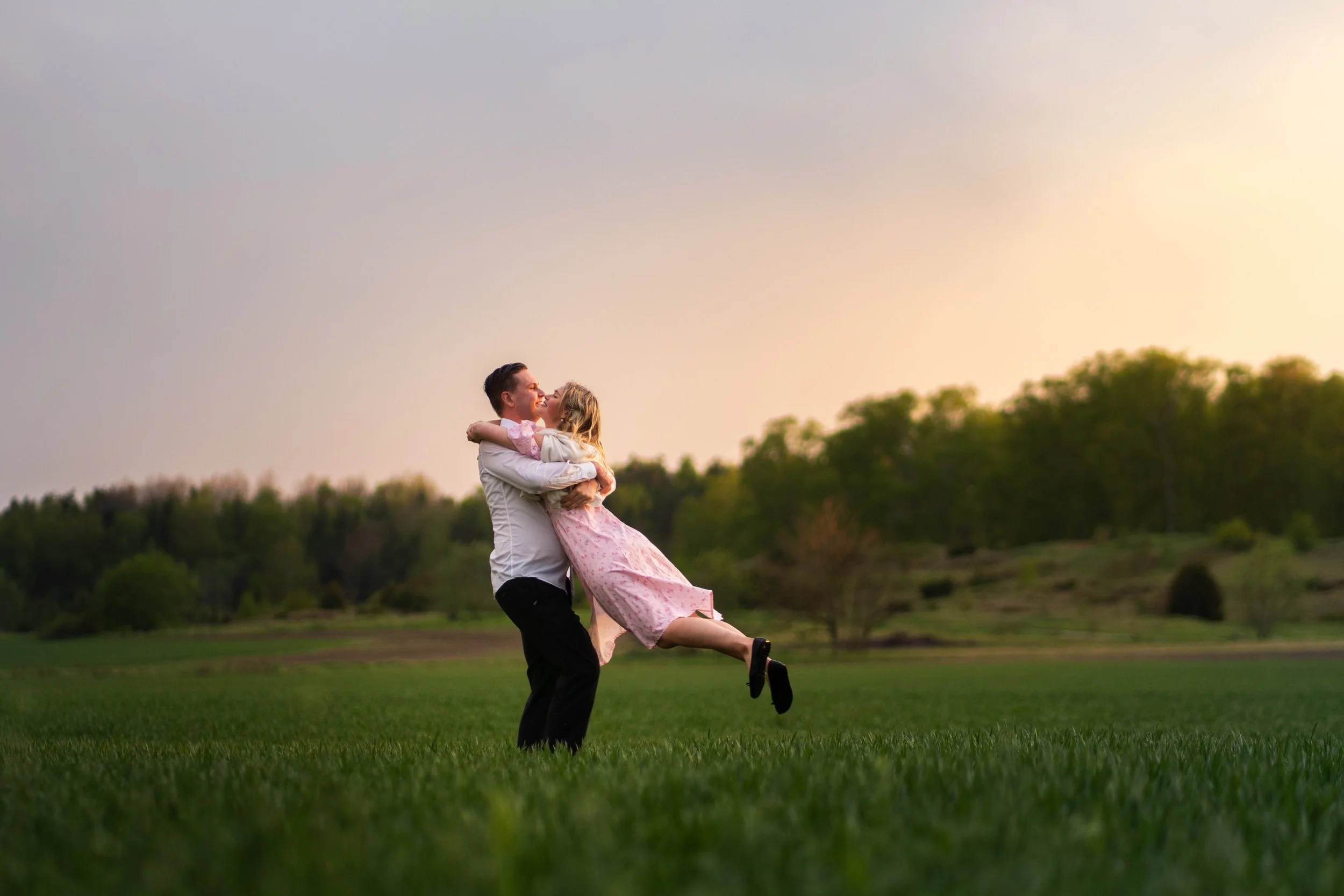 A man lifting a woman in a pink dress in an open grassy field during sunset.