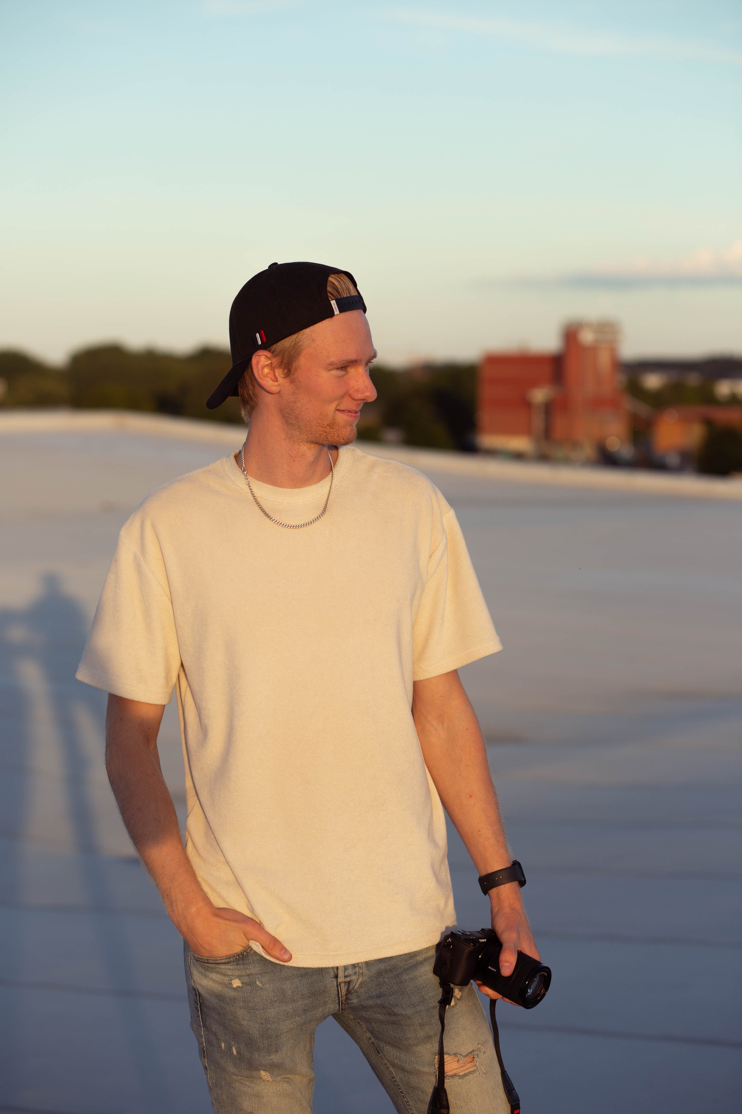 A young man with red hair wearing a black baseball cap backwards, a cream T-shirt, ripped jeans, and a silver chain necklace. He is holding a camera in his right hand and looking to his right with a slight smile. The background shows a sky during sunset or sunrise and some buildings in the distance.