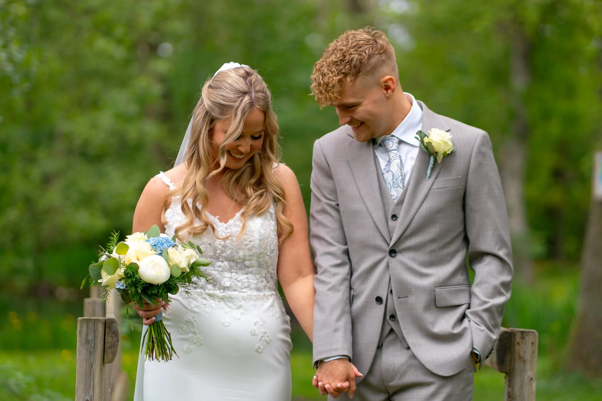 A newlywed couple holding hands and smiling in a green outdoor setting during their wedding.