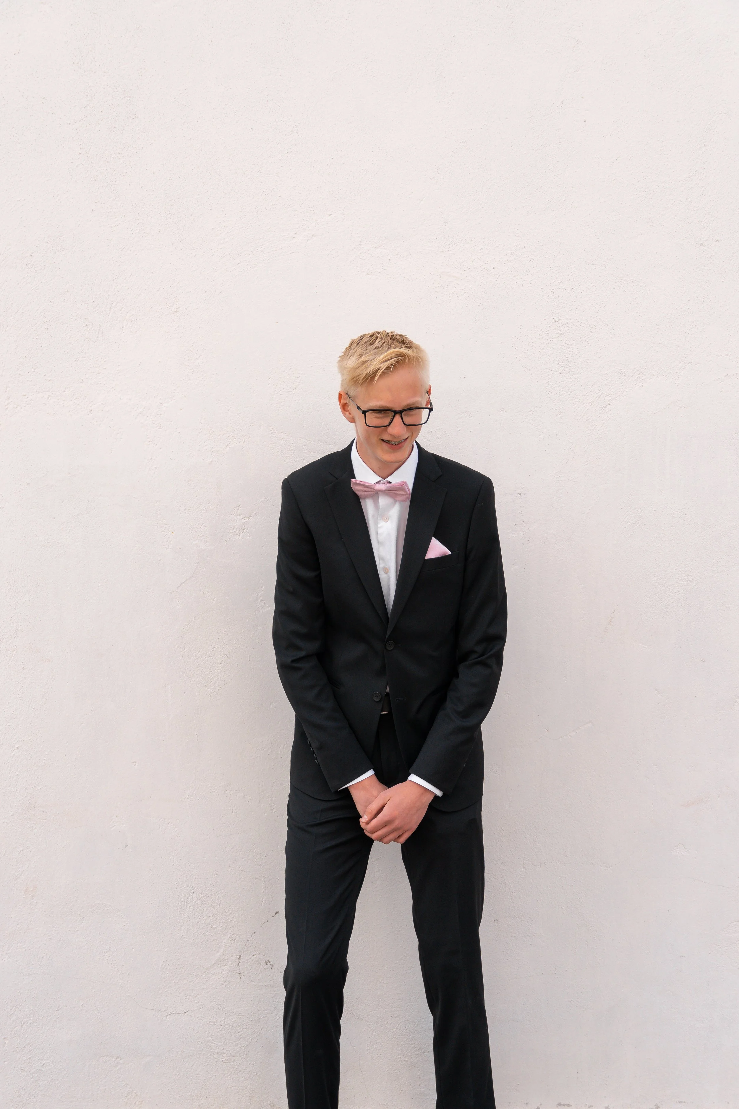 A young man in a black suit with a pink bow tie and pocket square, standing against a plain white wall, smiling with his hands clasped in front of him.