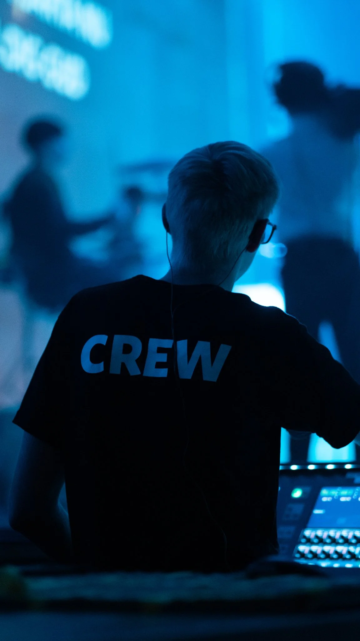 Person med tröja med texten 'CREW' som arbetar vid en ljud- eller ljusutrustning i ett mörkt utrymme, antagligen under en livehändelse eller produktion.