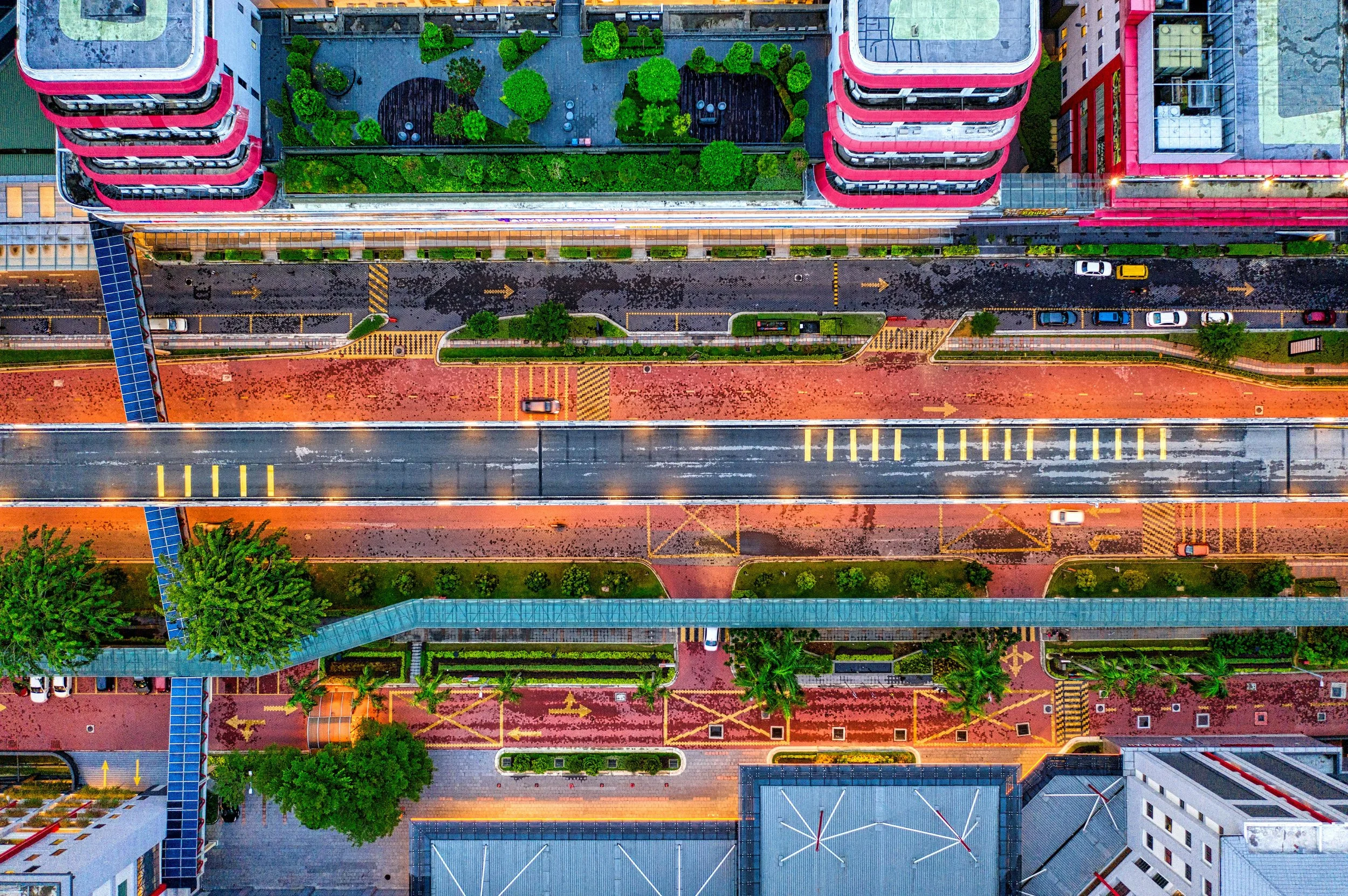 An aerial view of a city street at night, showing cars, crosswalks, sidewalks, trees, and surrounding buildings with green spaces and rooftop gardens.