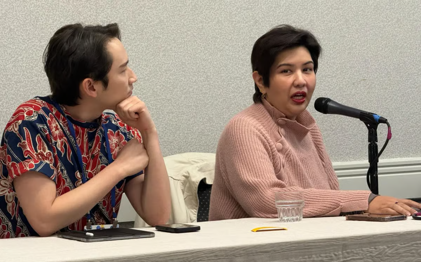 A man and woman are sitting at a table during a discussion or presentation, with the woman speaking into a microphone and the man listening attentively.
