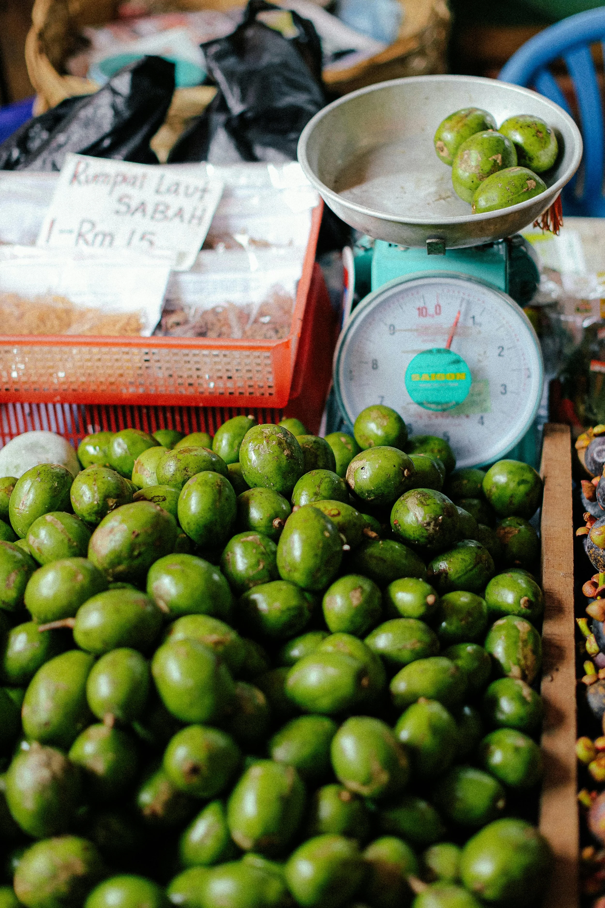 A market stall selling fresh green local fruits, likely lemon or lime, with a weighing scale and a plastic basket in the background with a sign showing prices.