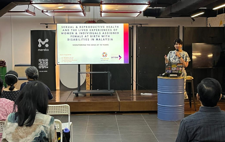 A woman speaking at a presentation on sexual and reproductive health and lived experiences of women with disabilities in Malaysia, with an audience seated in front of her, a large screen displaying the presentation title, and banners on the side.