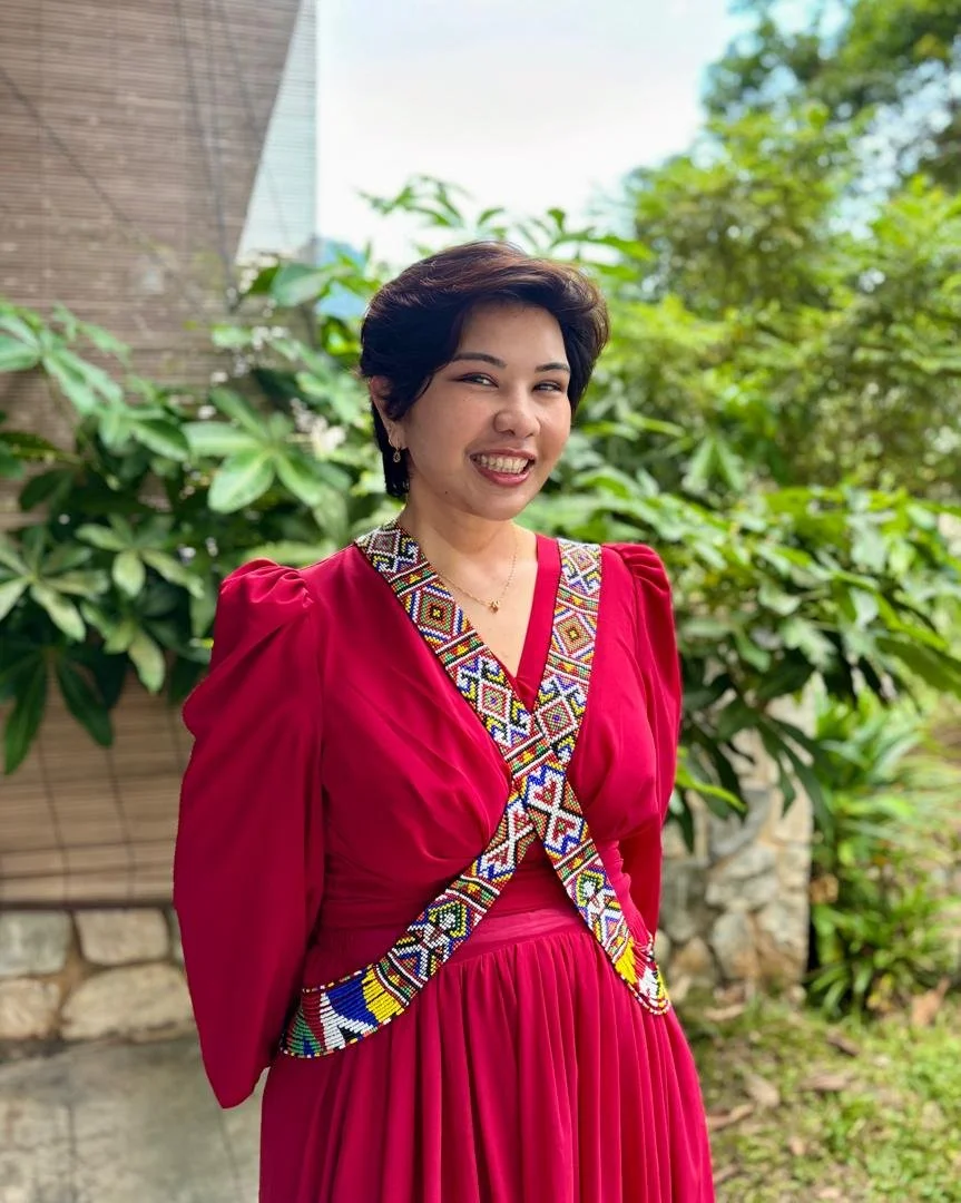 A woman with short dark hair wearing a vibrant red dress with colorful embroidered trim, standing outdoors in front of lush green foliage and a brick wall.