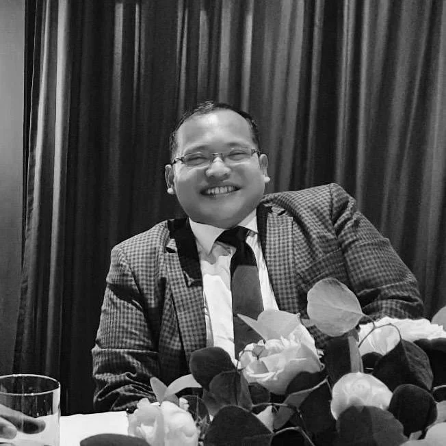 A smiling man with glasses in a suit and tie, sitting at a table decorated with flowers, in front of a dark curtain.