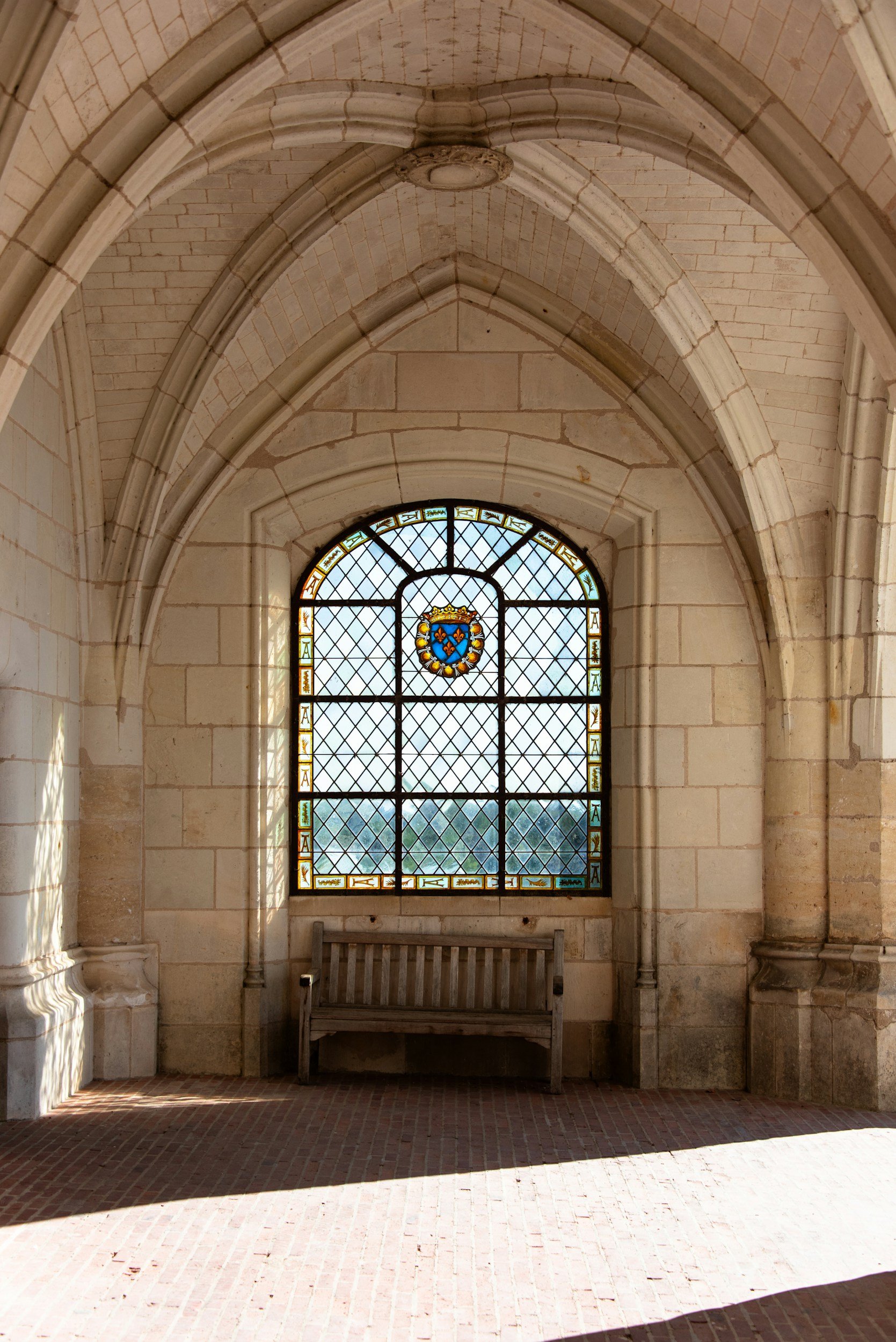 A stained glass window with a coat of arms and a view of a landscape outside, inside a stone building with a bench beneath the window and Gothic arches overhead.