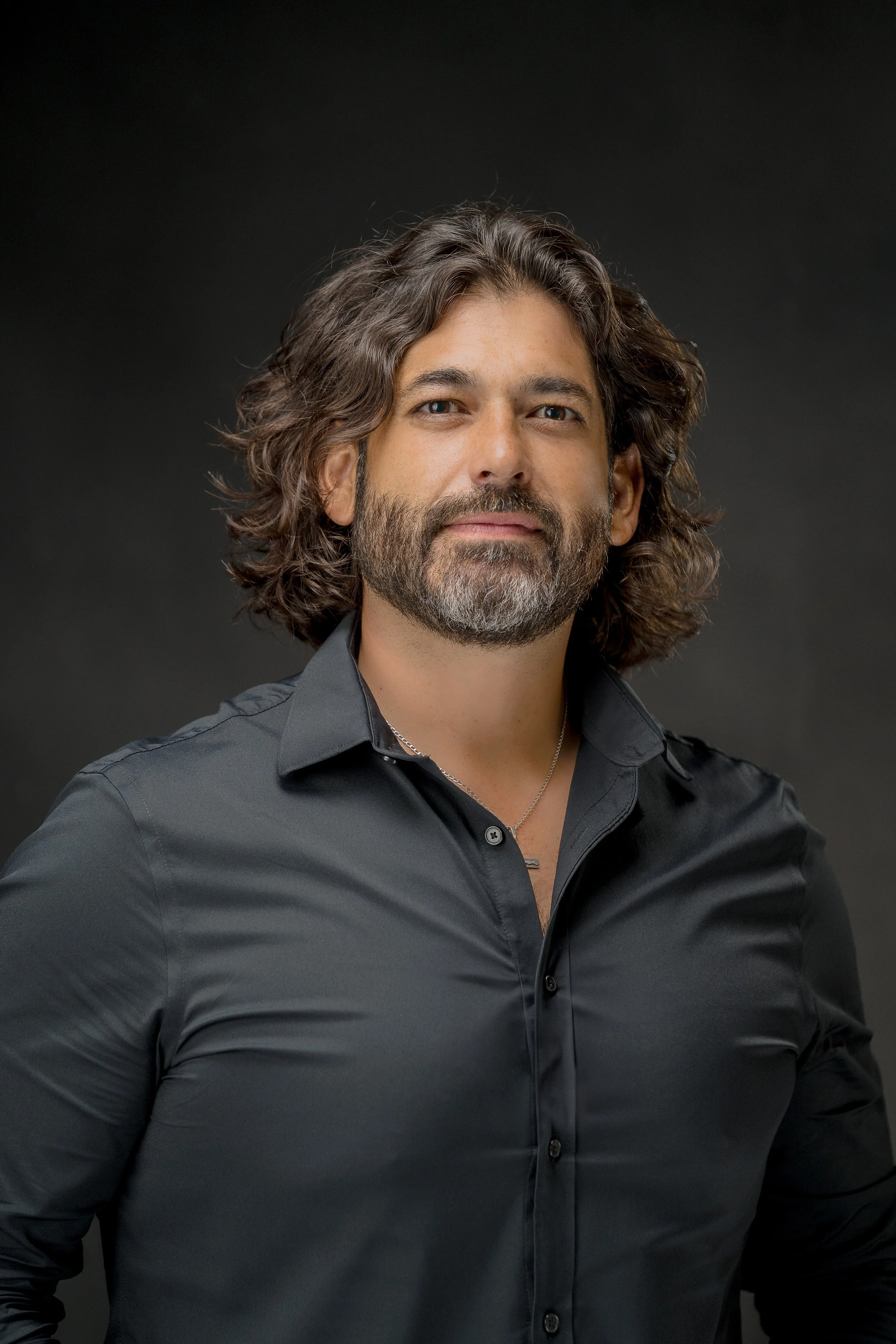 Portrait of a man with long, curly hair and a beard, wearing a black button-up shirt, standing against a dark background.
