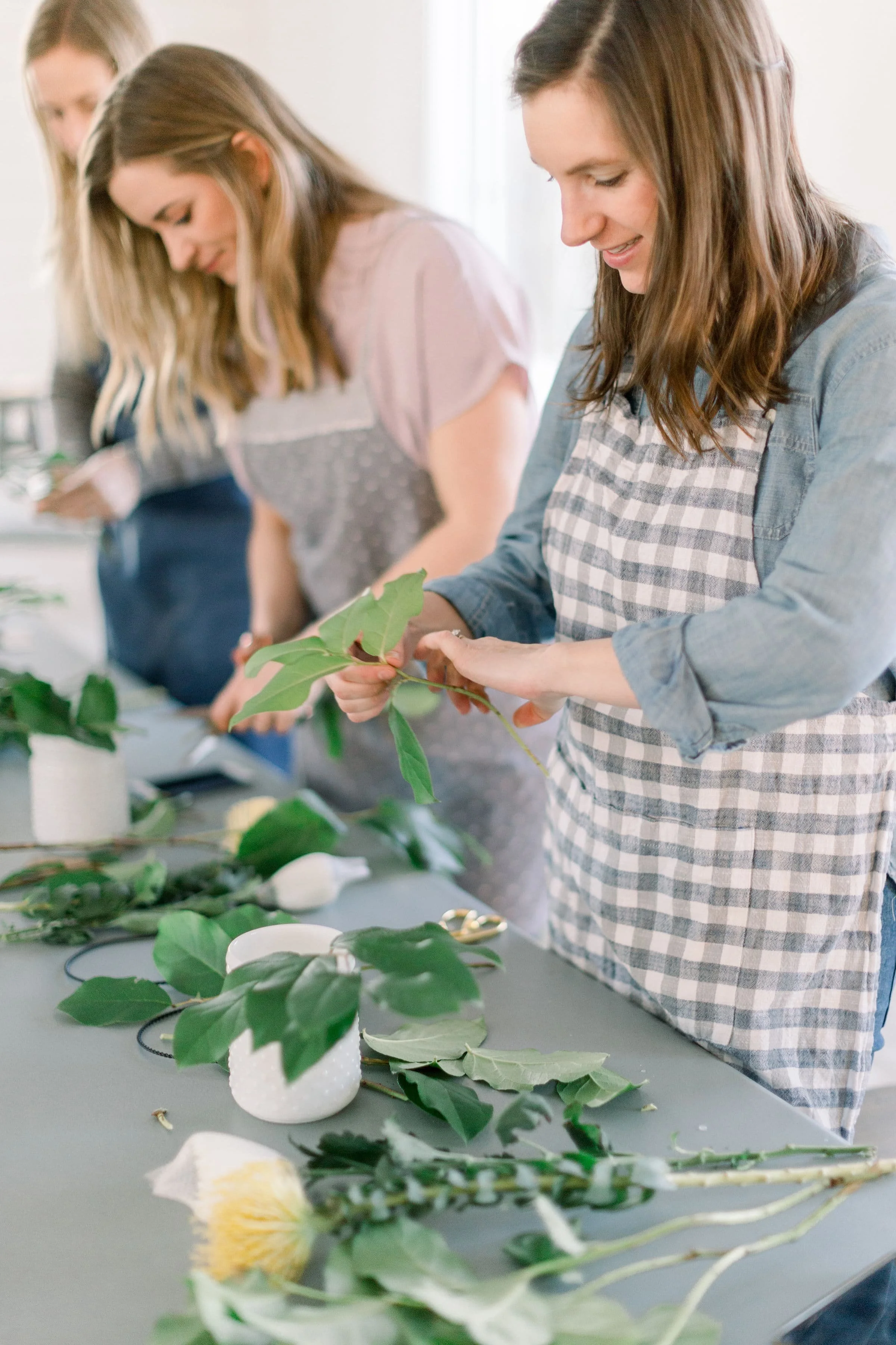 A group of ladies wearing aprons, standing at a table. They are cutting greenery and flowers to make an arrangement at a Twig & Bloom flower workshop in Edmonton, AB.