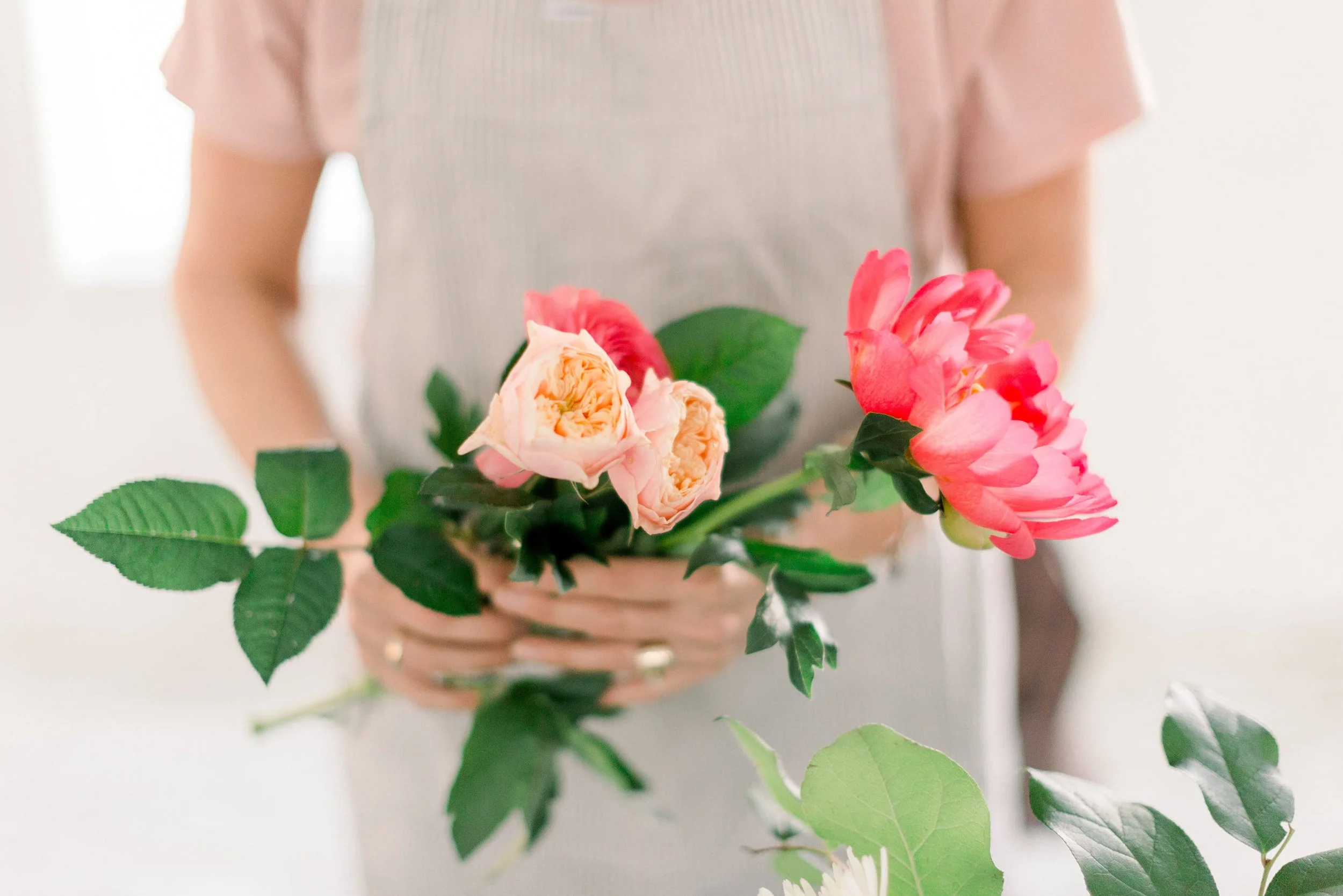 A young women wearing an apron holding some coral peonies and peach roses at a Twig & Bloom workshop in Edmonton, AB.