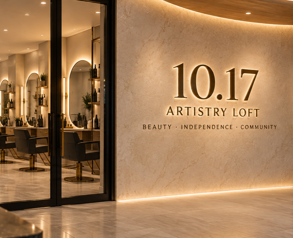 Interior of a beauty salon with black chairs, mirrors, and shelves with products, along with illuminated wall signage that reads '10.17 Artistry Loft' and words 'Beauty, Independence, Community'.