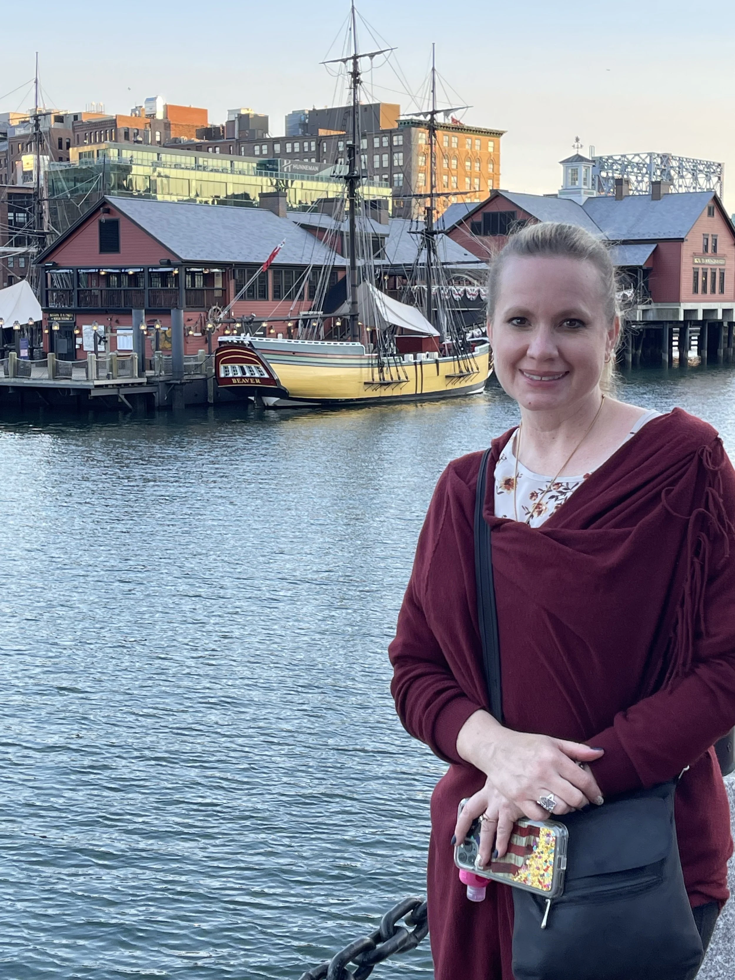 A smiling woman standing near a waterfront with boats, including a large yellow sailing ship, and historic buildings in the background.