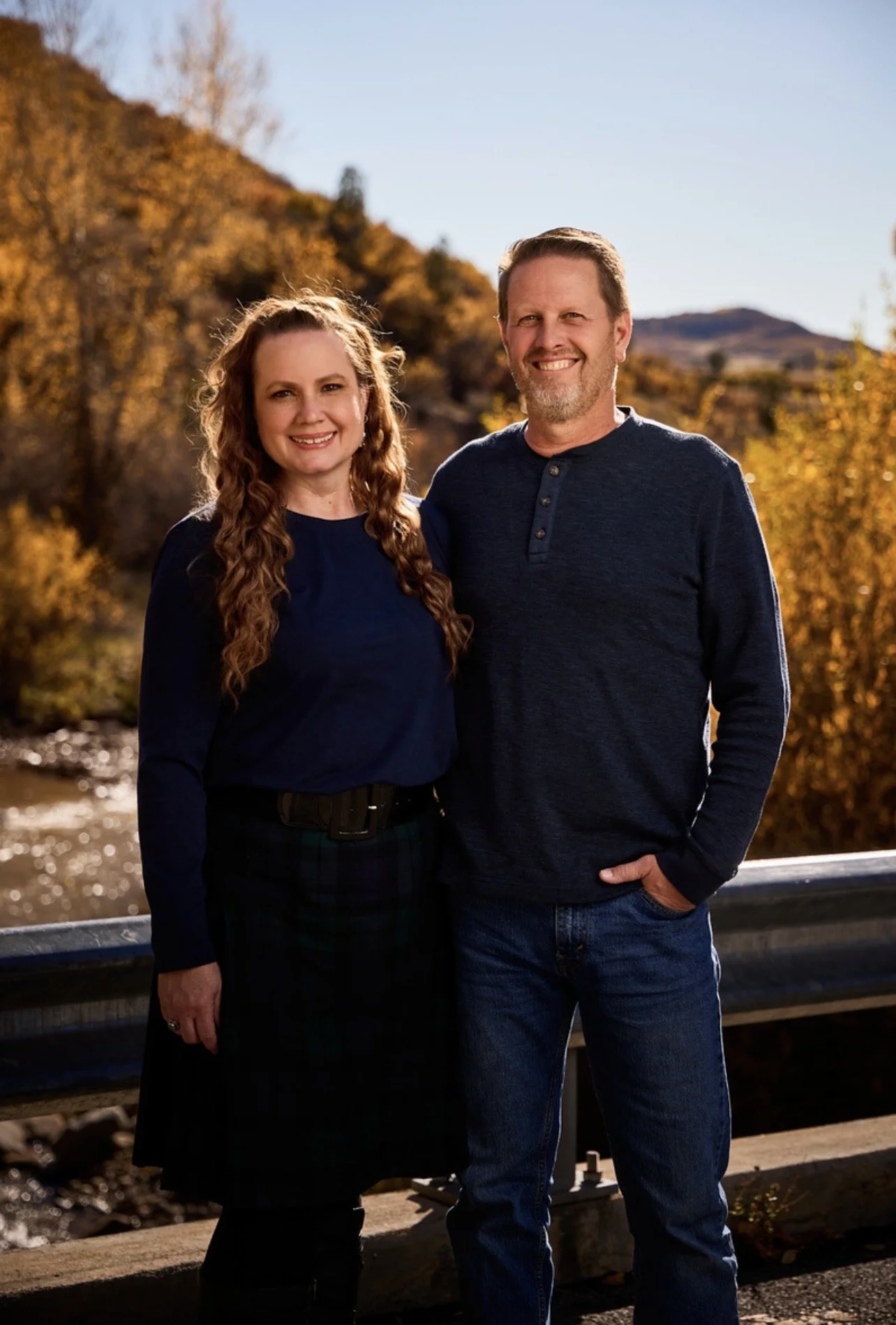 A smiling man and woman standing outdoors on a bridge in autumn, with orange and yellow trees and a mountain in the background.