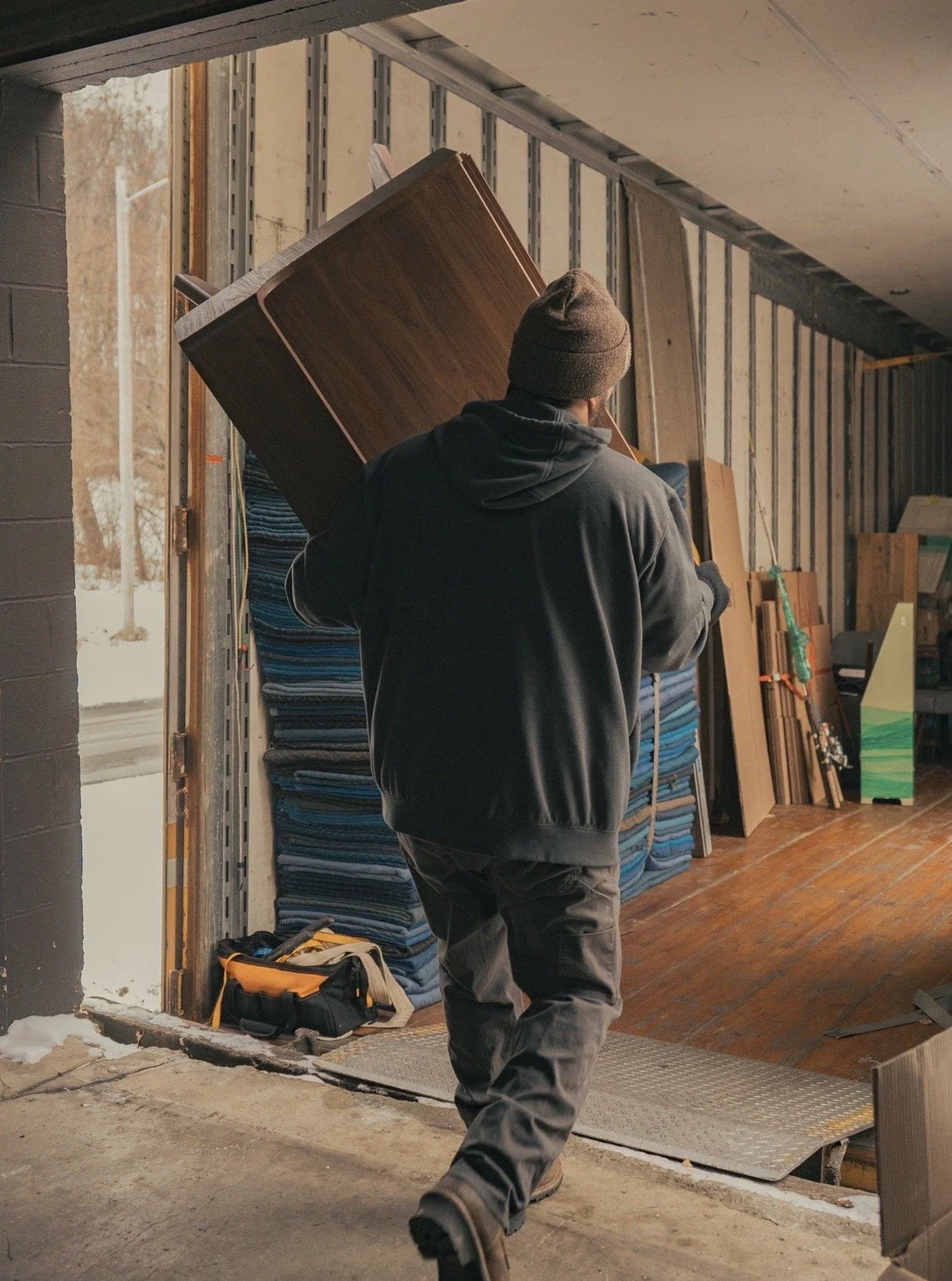 A person in dark clothing and a beanie carrying a wooden table into a building under construction or renovation, with building materials and tools around.