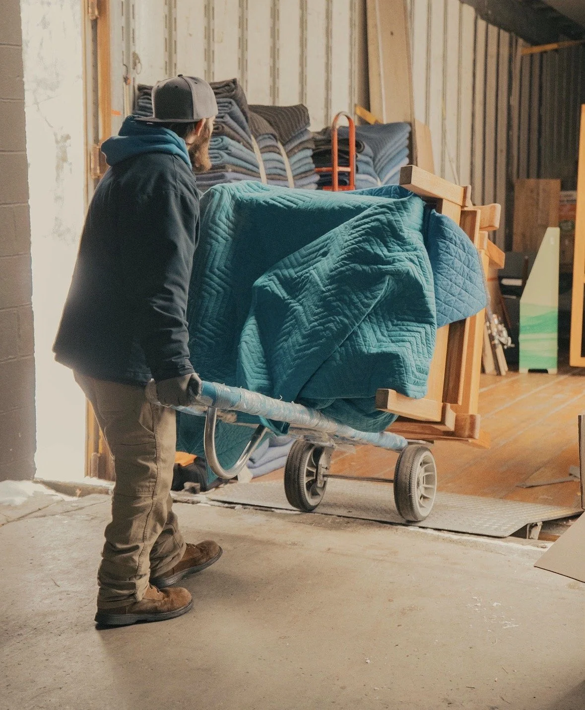 A person in work clothes and a baseball cap is pushing a cart loaded with furniture wrapped in a blue quilt inside a warehouse or storage area.