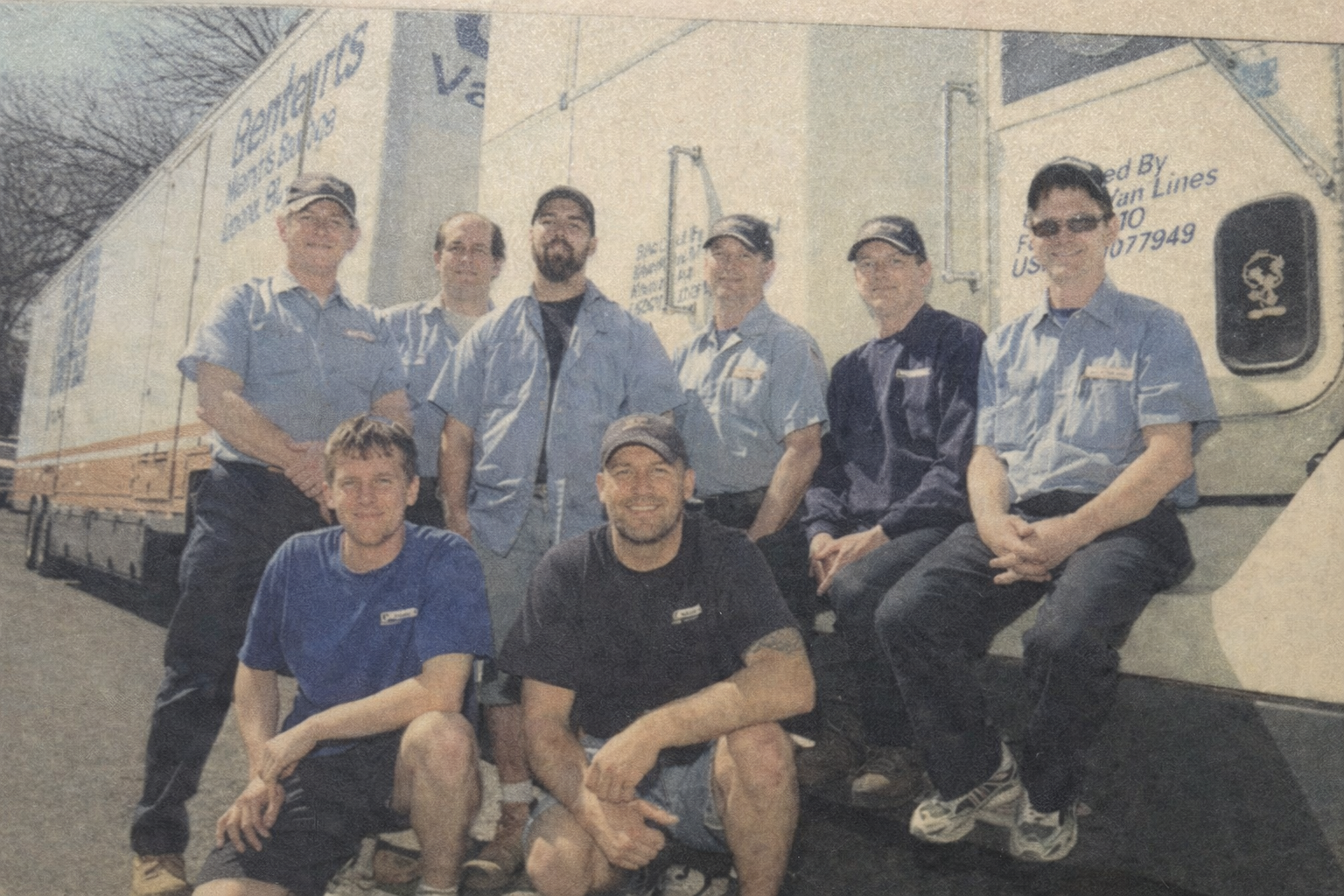 Group of eight men posing outdoors beside a large moving truck.