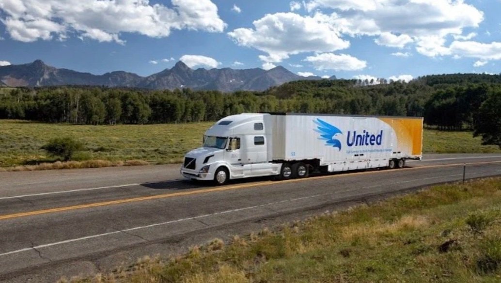 A white semi-truck with a colorful trailer driving on a highway amid green fields, trees, and mountains in the background under a partly cloudy sky.
