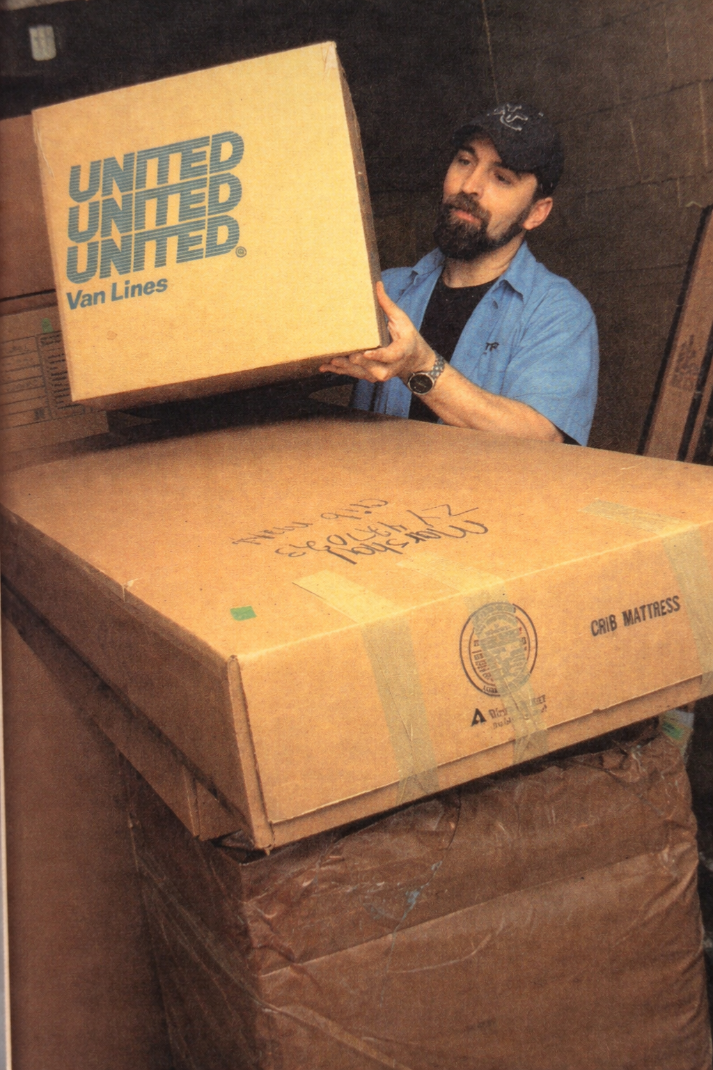 A man with a beard and dark hair wearing a black cap, a blue shirt, and a watch is lifting a cardboard box labeled "UNITED Van Lines" in a storage room filled with stacked boxes.