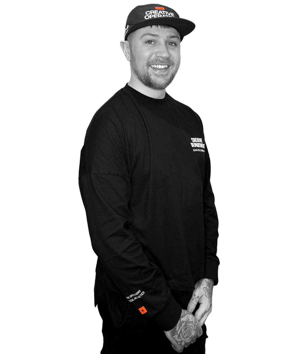 Joe Lane, brand designer wearing a black cap with white text and a black long-sleeve shirt with white text, smiling and standing with his hands clasped in front of him, revealing tattoos on his fingers and left hand.