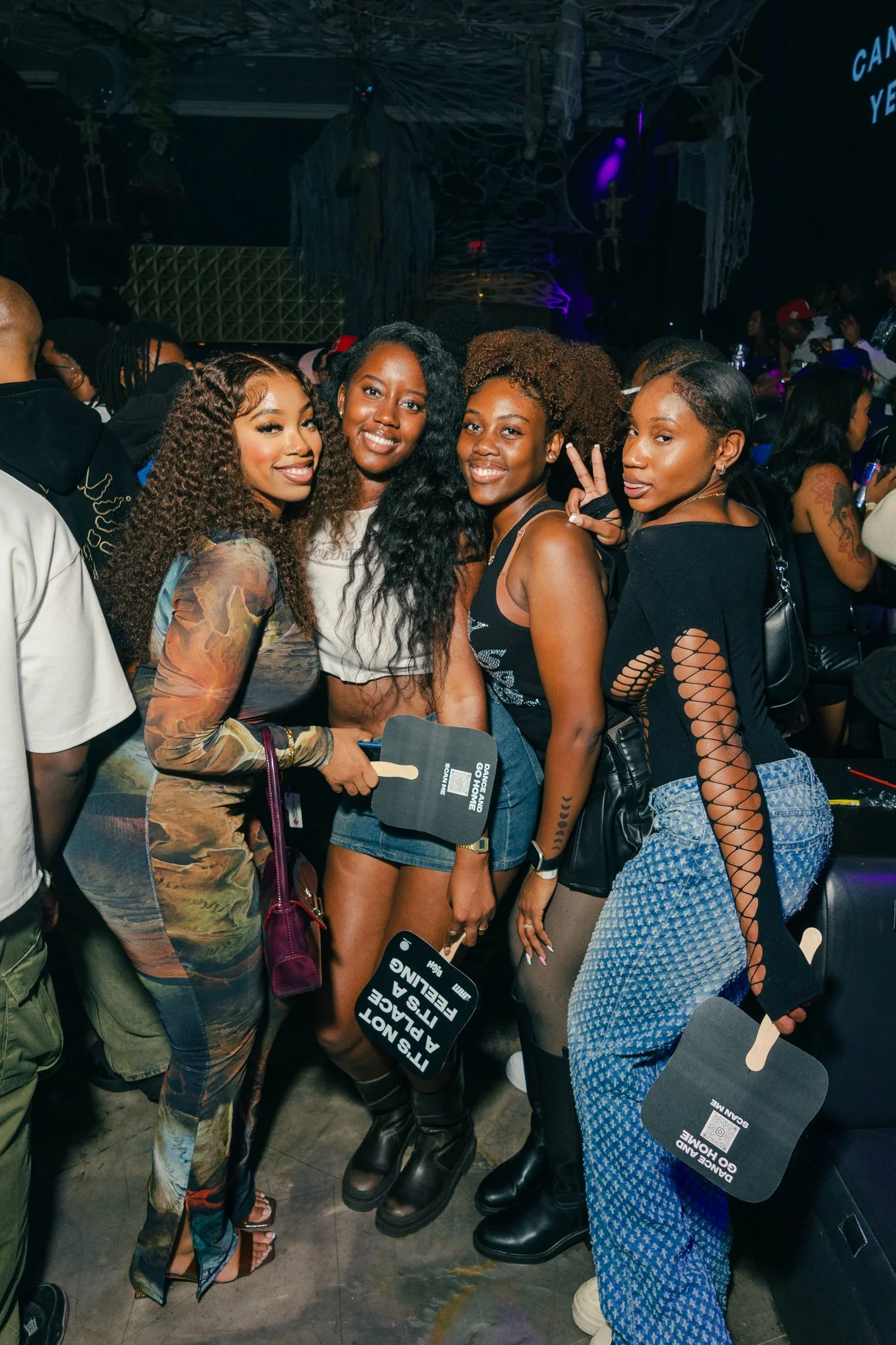 Four women posing together at a lively event or party, with decorations and other people in the background.