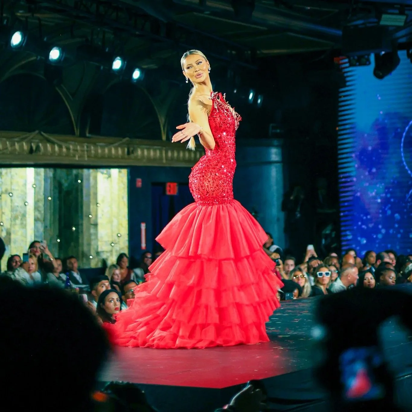 A woman on a stage wearing a red sequined mermaid gown with tiered ruffle skirt, striking a pose with her hand extended.
