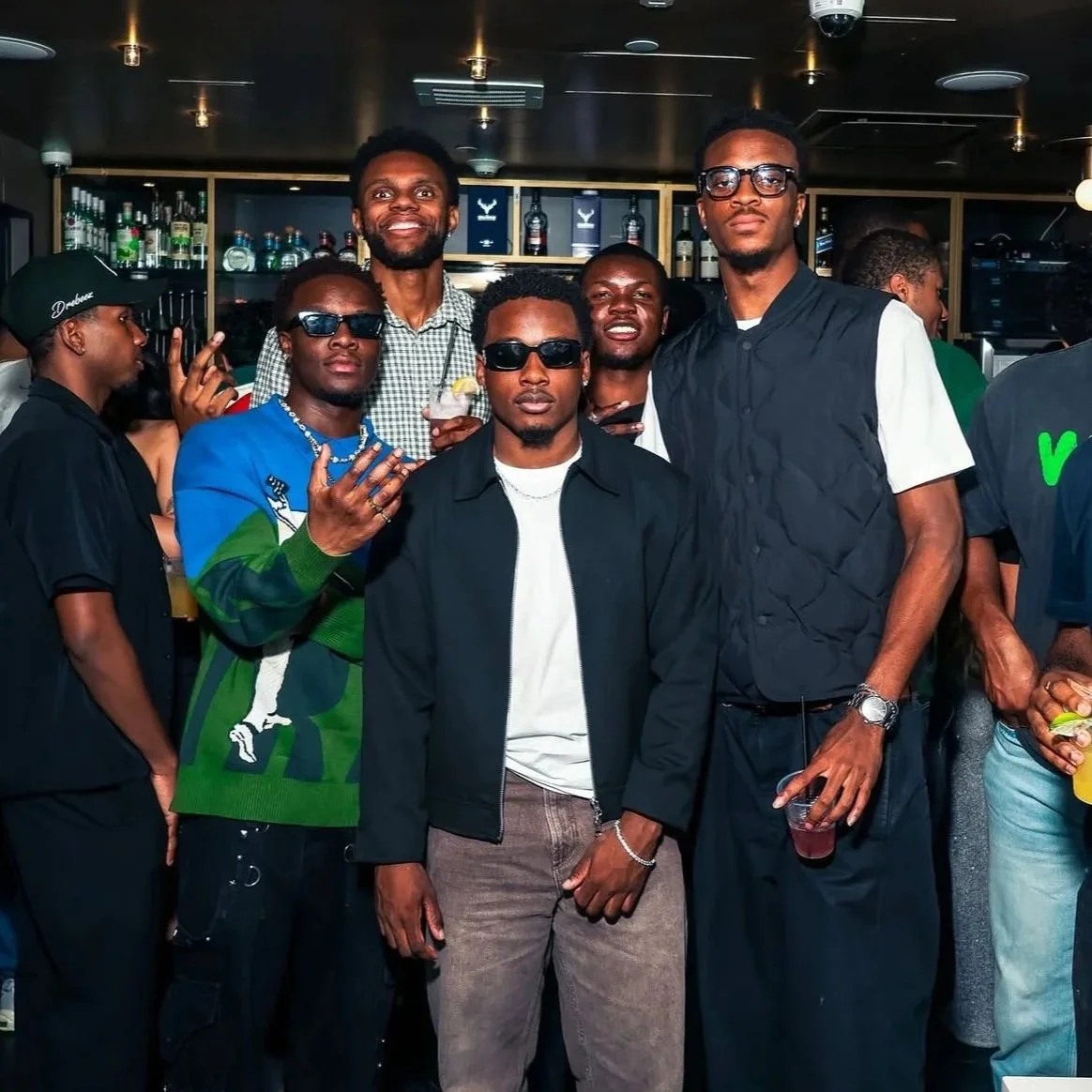 A group of men at a bar or nightclub, some wearing sunglasses and casual clothing, standing together and posing for the photo.