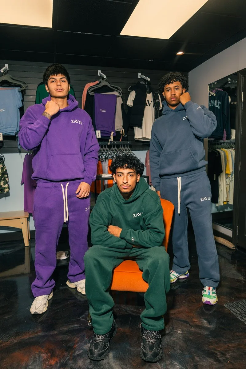 Three young men in colorful ZAVII hoodies and sweatpants inside a clothing store, with racks and shelves of apparel behind them.