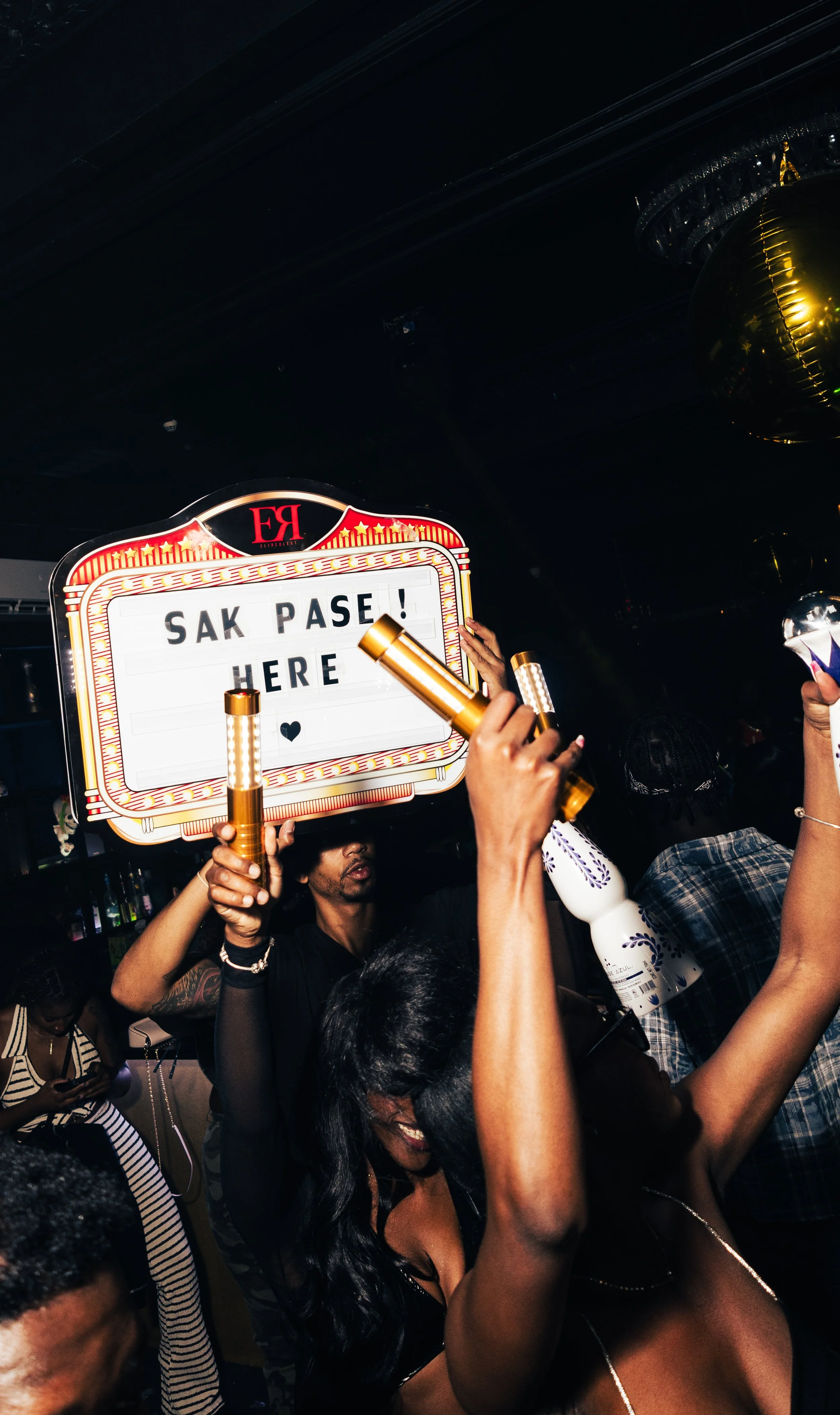 A group of people at a party holding a sign that reads 'SAK PASE! HERE' in a dimly lit setting with balloons.