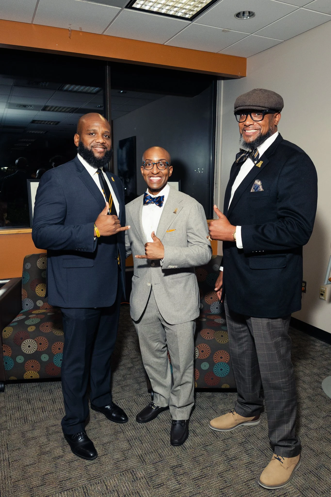 Three men dressed in suits and bowties standing indoors, smiling and making hand gestures for the camera.