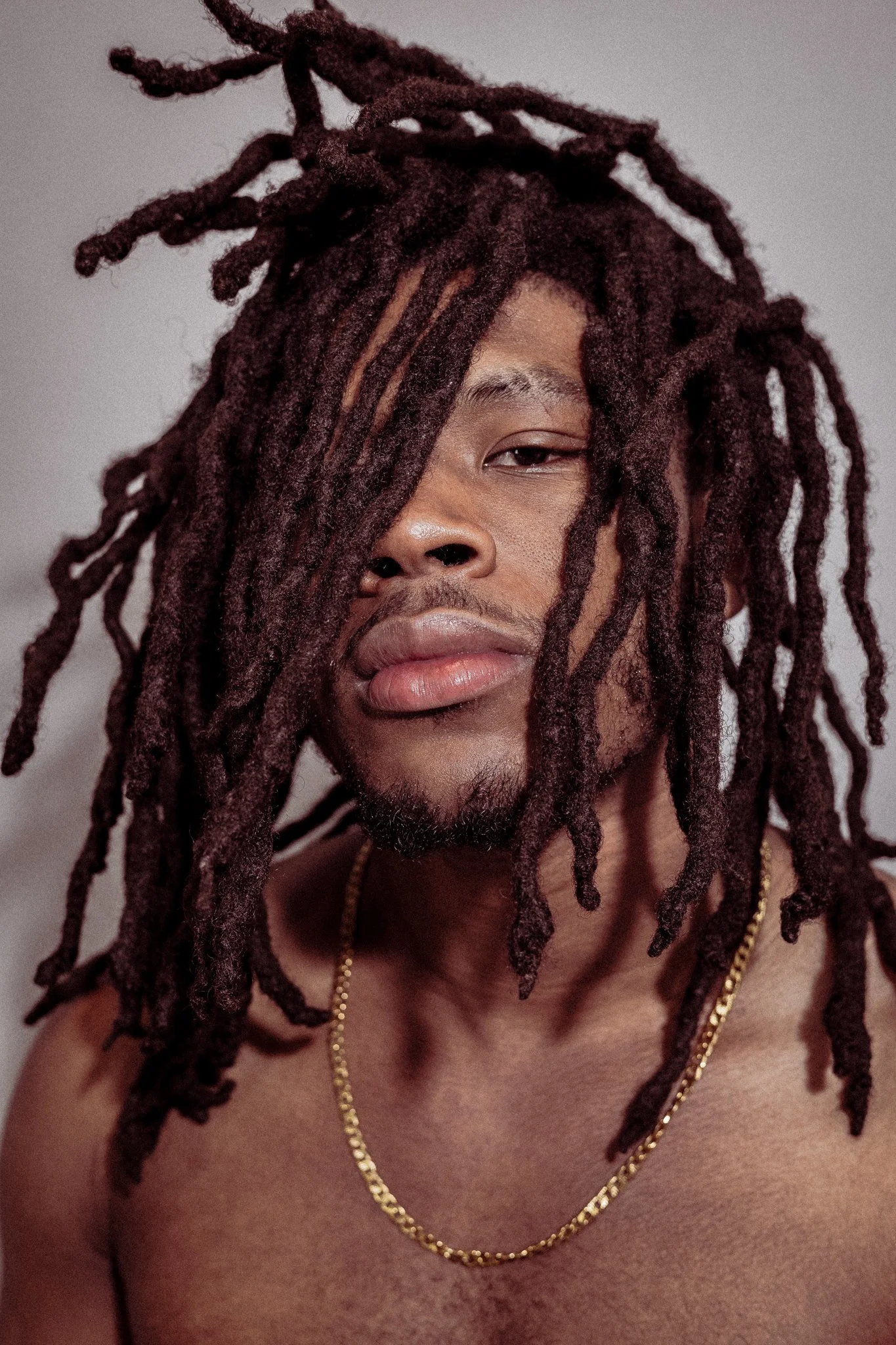A shirtless man with long, dreadlocked hair, wearing a gold chain necklace, with a neutral expression.