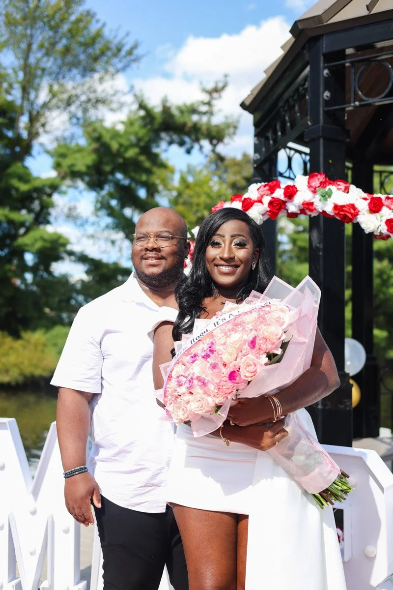 A smiling couple at a wedding celebration, with the woman holding a bouquet of pink and white roses, standing outdoors near floral decorations and a black metal structure with red and white flowers.