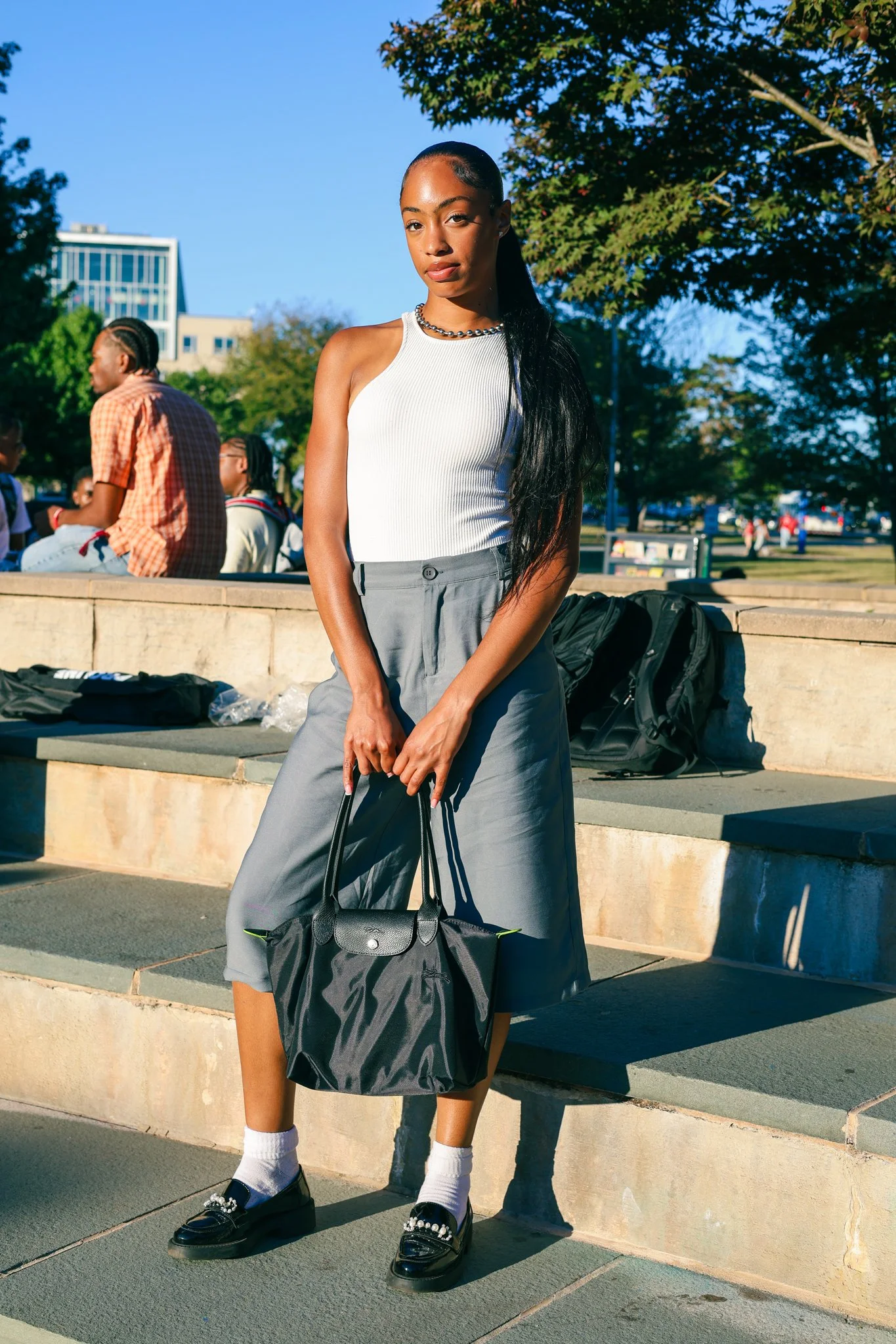 A young woman with a high ponytail wearing a sleeveless white top, gray wide-leg pants, black loafers with embellishments, and white socks, holding a black handbag, standing outdoors on steps with a park and other people in the background on a sunny day.