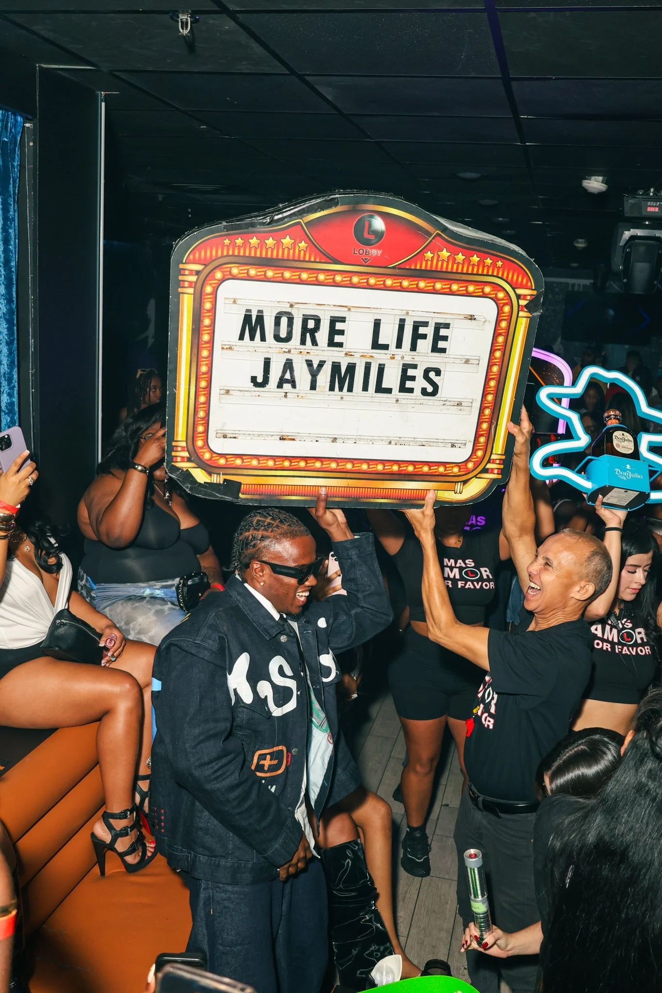 Two people hold a large illuminated sign that reads "MORE LIFE JAYMILES" in a crowded bar or nightclub scene. The person on the left is wearing sunglasses and a dark jacket, smiling, while the person on the right is wearing a black shirt and also smi