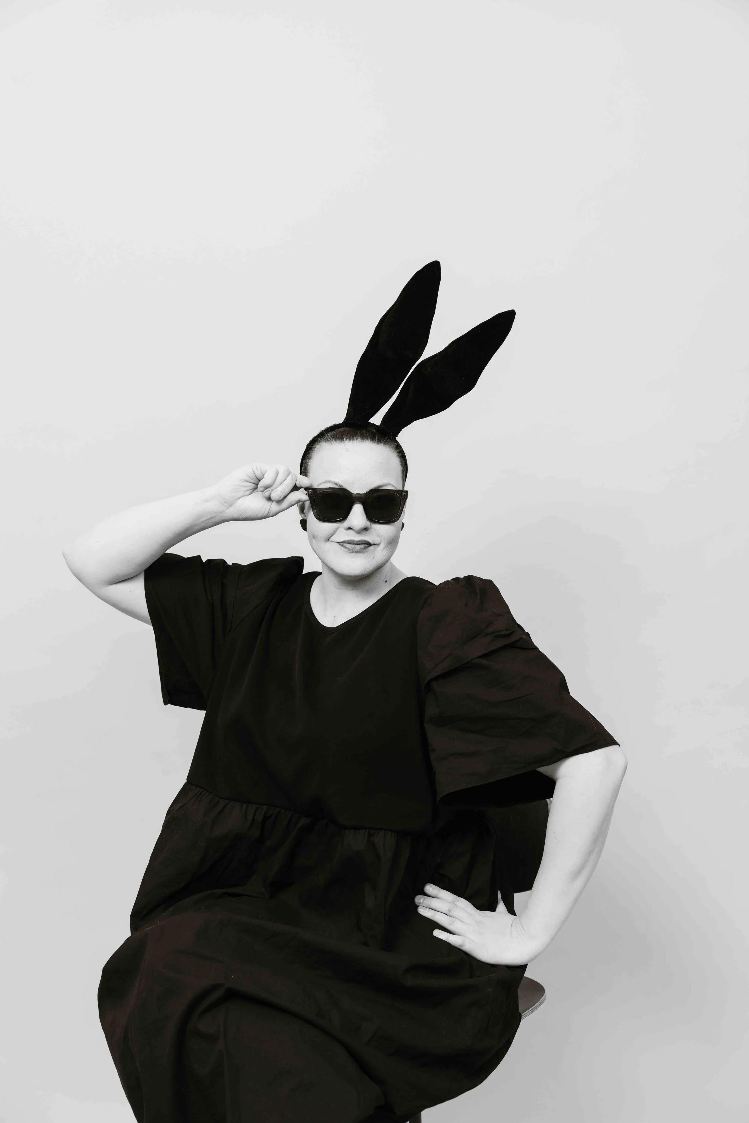 Nainen pukeutuneena mustaan mekkoon ja bunny-hattuun, pitää kädellään silmälaseja ja istuu tuolilla, mustavalkokuva.
