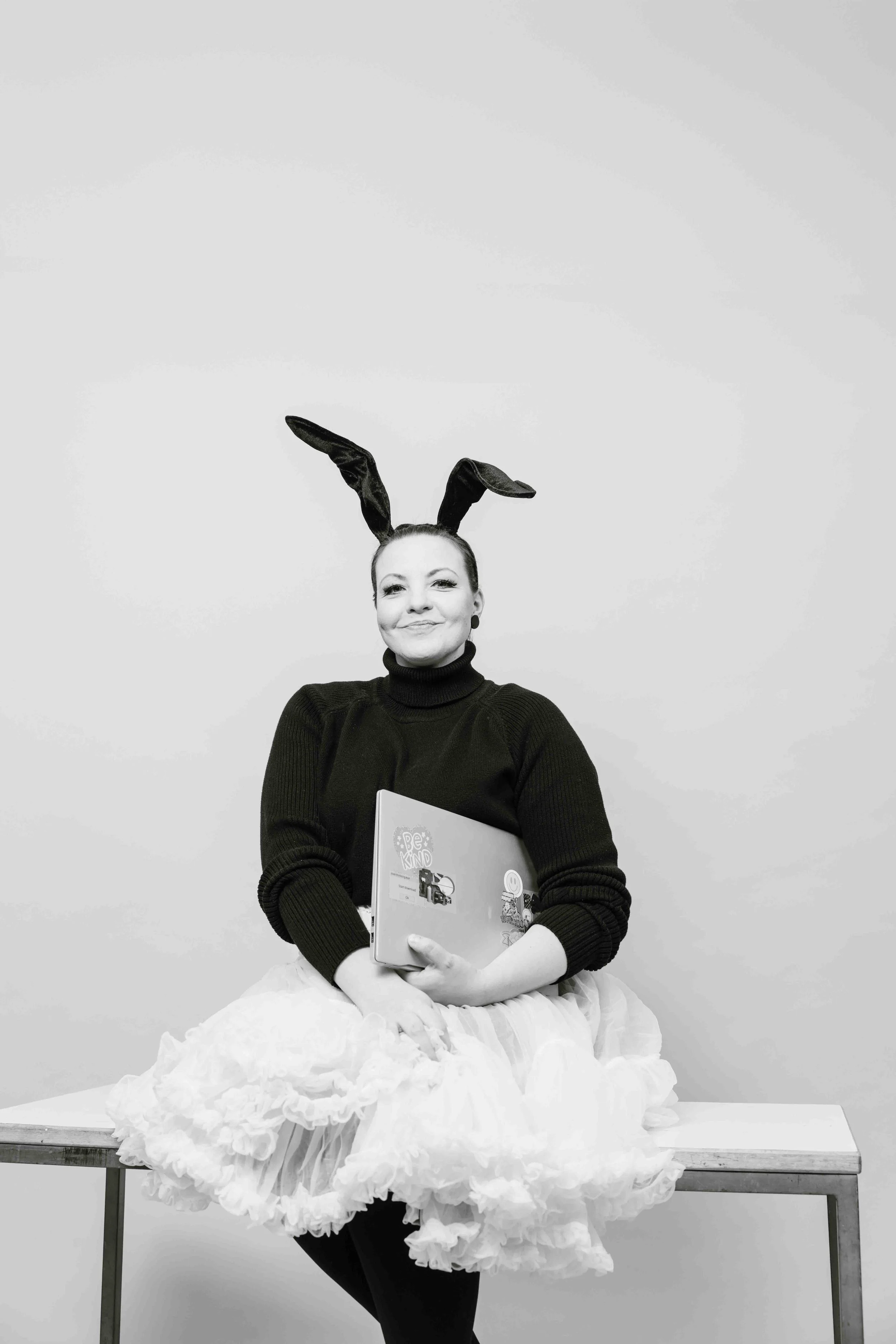 Nainen istuu pöydän päällä, hänellä on pupun korvat ja röyhelöinen hame, ja hän pitää käsissään kannettavaa tietokonetta.