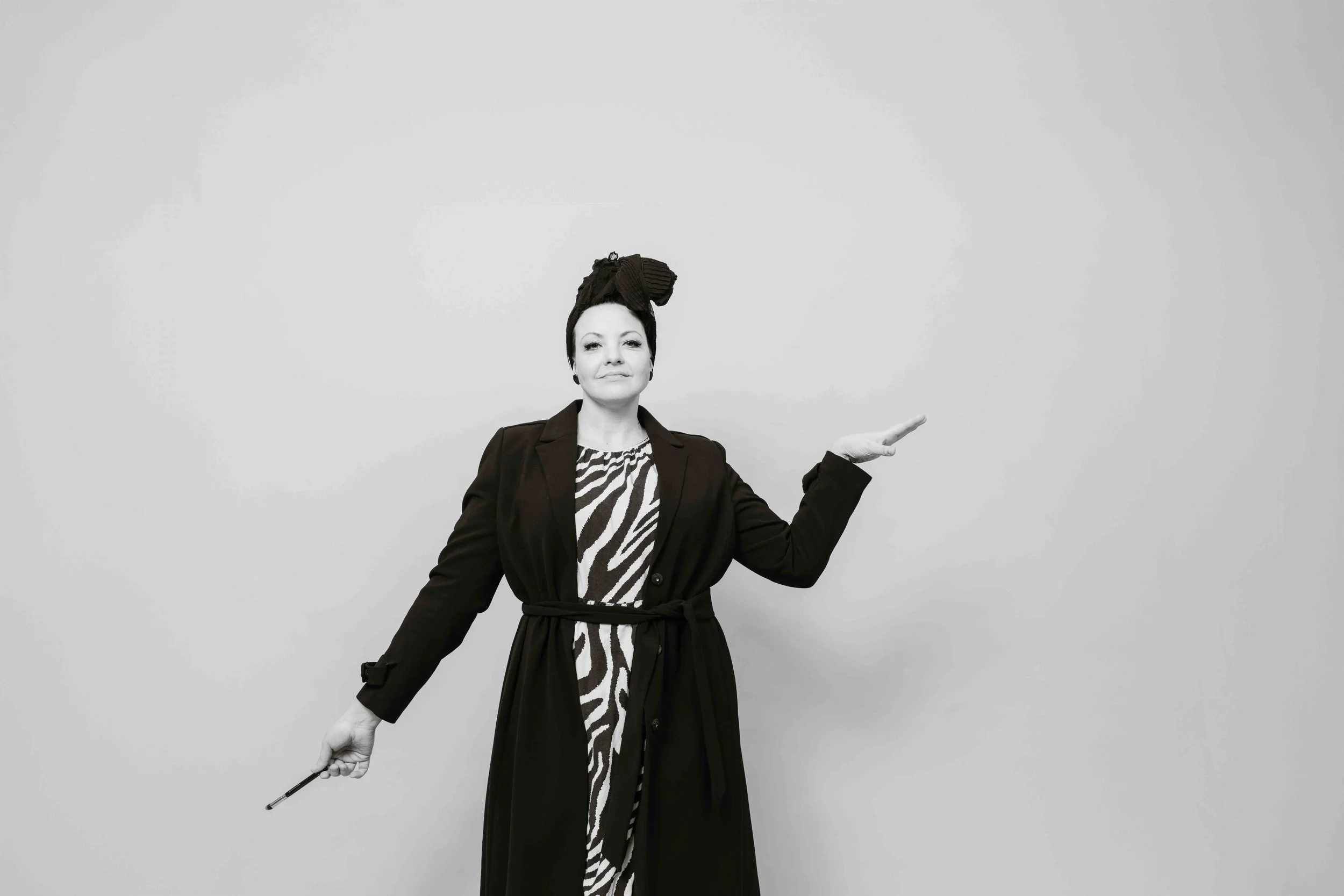 Mustavalkoinen kuva naisesta, jolla on huivi pään ympärillä, yllään zebra-kuvioinen mekko ja musta panta, seisoo tyhjän valkoisen seinän edessä ja pitää kädessään taikasauvaa.