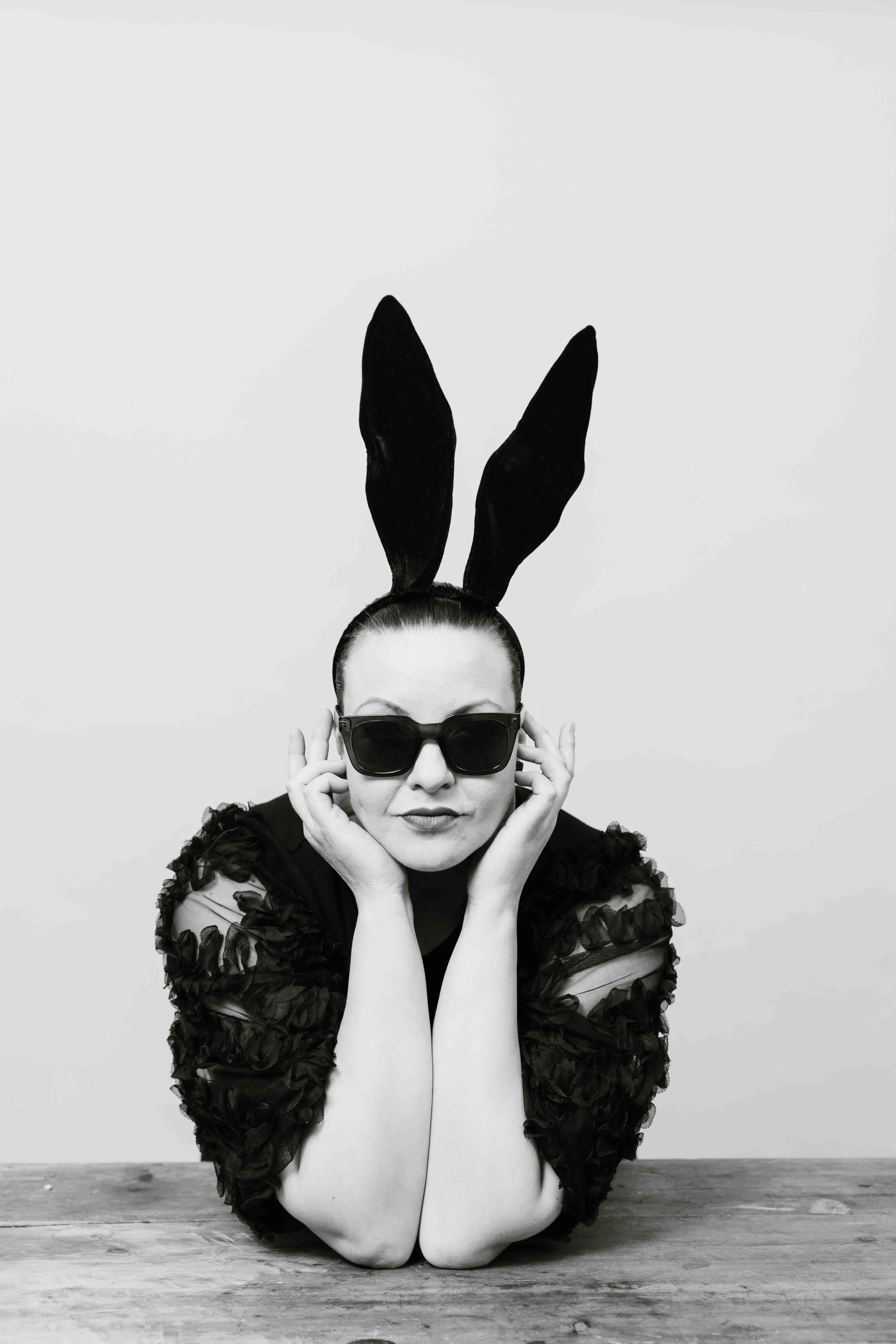 Nainen, jolla on suuret mustat silmälasit ja mustat pupunkorvat, istuu pöydän ääressä, katsoo kameraan.