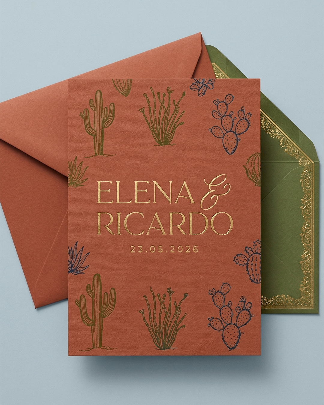 Tarjeta de invitación de boda con fondo color terracota, decorada con dibujos de cactus y suculentas en azul y verde, con nombres Elena y Ricardo en letras doradas y fecha 23.05.2026, acompañada de sobres verde con borde dorado y rosa con borde dorad