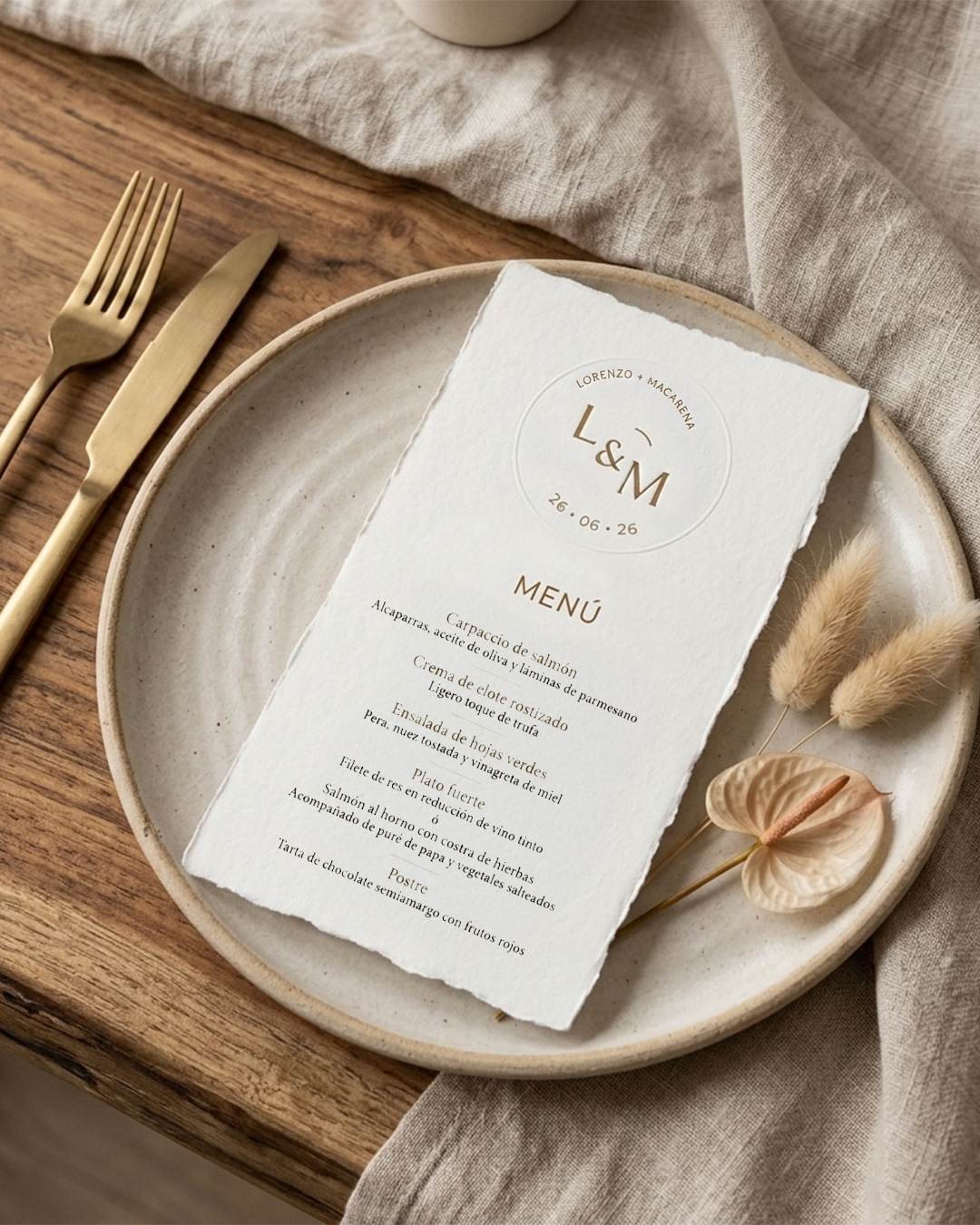 Menú de boda en papel con borde irregular, sobre un plato de cerámica, acompañado de cubiertos dorados y decoración con flores secas, en una mesa de madera con tela beige.