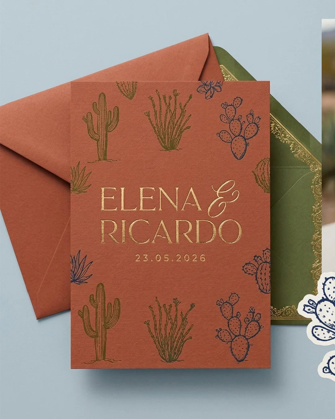 Tarjeta de invitación de boda con fondo color terracota y dibujos de cactus y plantas suculentas, con los nombres Elena y Ricardo y la fecha 23.05.2026, acompañada de sobres verdes y decoraciones relacionadas con el tema del desierto.