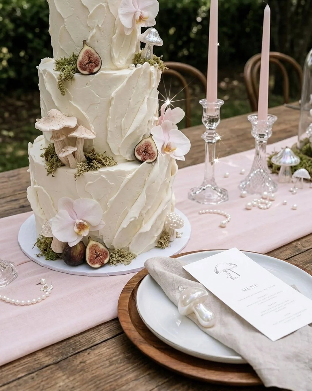 Pastel de boda con decoración de hongos, flores y frutas sobre mesa de madera, con candelabros y velas rosadas.
